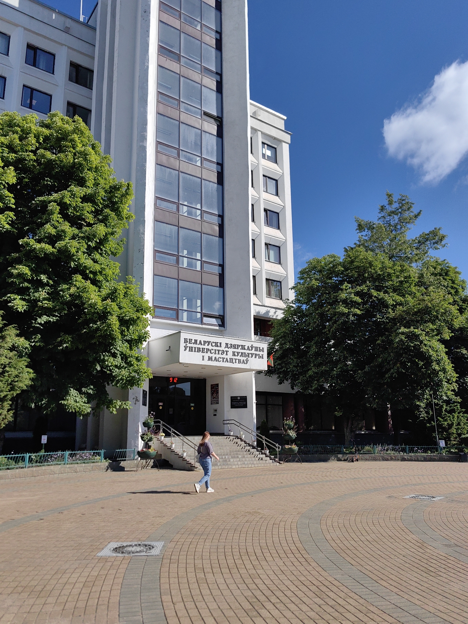 This is an eye-level, medium shot of the Belarusian State University of Culture and Arts, located in Minsk, Belarus. The university building is a tall, white structure with a prominent glass facade. In front of the building, there is a paved courtyard with a circular pattern. A young woman in jeans and a purple shirt is walking across the courtyard from right to left. Lush green trees flank the building on both sides, and the sky above is a bright blue with a single white cloud. The overall scene depicts a sunny day on a university campus.