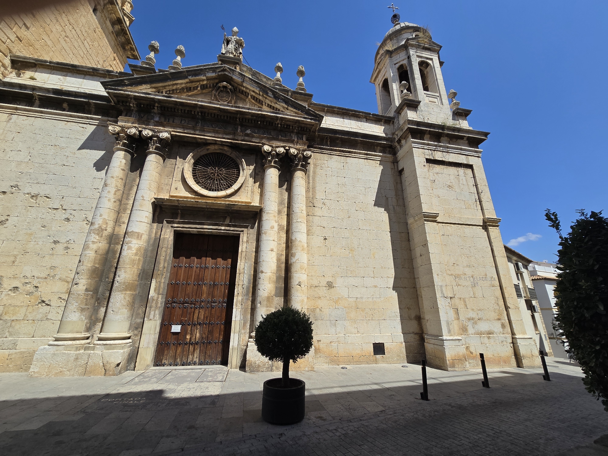 The image shows the facade of a stone church with a bell tower and a large wooden door. A small, round, potted tree stands in front of the door. The sky is a clear, bright blue, and sunlight casts sharp shadows on the ground. The architectural style suggests a historical building, possibly in a Mediterranean climate. The building appears to be located in the city of Jaén, Spain, as indicated by the inscription on the wall of the bell tower that reads "IVAN VERLACE, 1886". The street in front of the church is paved with stone tiles, and there are several black bollards along the side of the street. A portion of a dark green tree or bush is visible on the right side of the frame, partially obscuring buildings in the background.