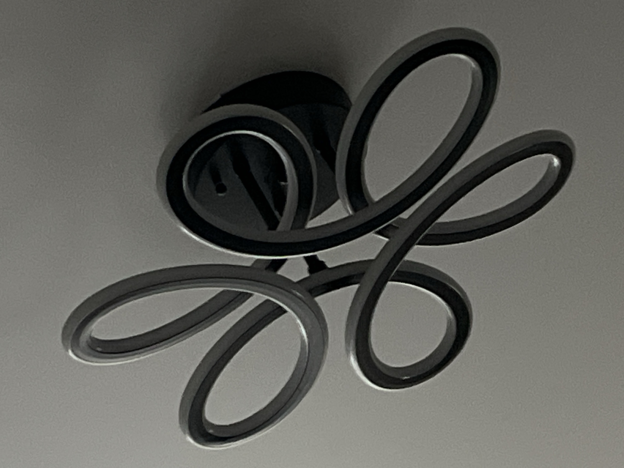 The image captures a modern ceiling light fixture from below, set against a light grey ceiling. The fixture is composed of multiple intertwined, ribbon-like elements that create an abstract, looping design. These elements feature a metallic or silver-toned outer edge and a darker, possibly recessed, inner surface where the light source (likely LED strips) would be located. The fixture is mounted to the ceiling by a central dark, circular base. The overall lighting in the photograph is subdued, giving the scene a muted, somewhat desaturated appearance. No specific activity or event is taking place, and there are no people or visible text. The image provides a close-up view of the fixture's contemporary design.