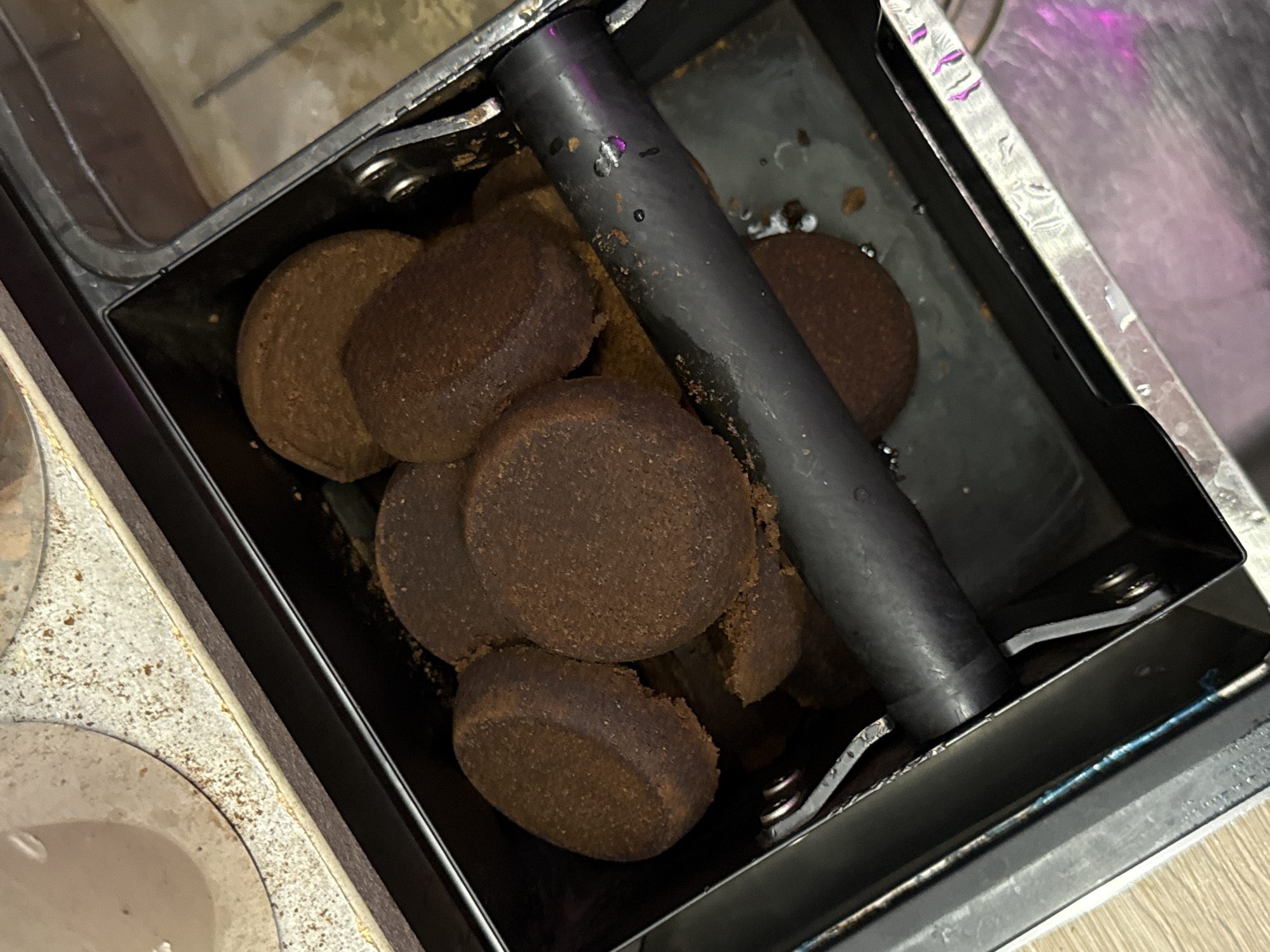 A close-up, high-angle shot captures a black metal waste bin filled with used coffee pucks. The pucks are dark brown and circular, with some appearing slightly broken. A black cylindrical bar rests horizontally across the pucks. The interior of the bin shows some moisture and small brown particles. The bin itself is set within a larger stainless steel structure, which appears to be part of a coffee machine. The surrounding environment is out of focus, but hints of a light-colored speckled countertop and a pinkish glow suggest an indoor setting. No specific location in Moscow, Russia, can be identified from this image.