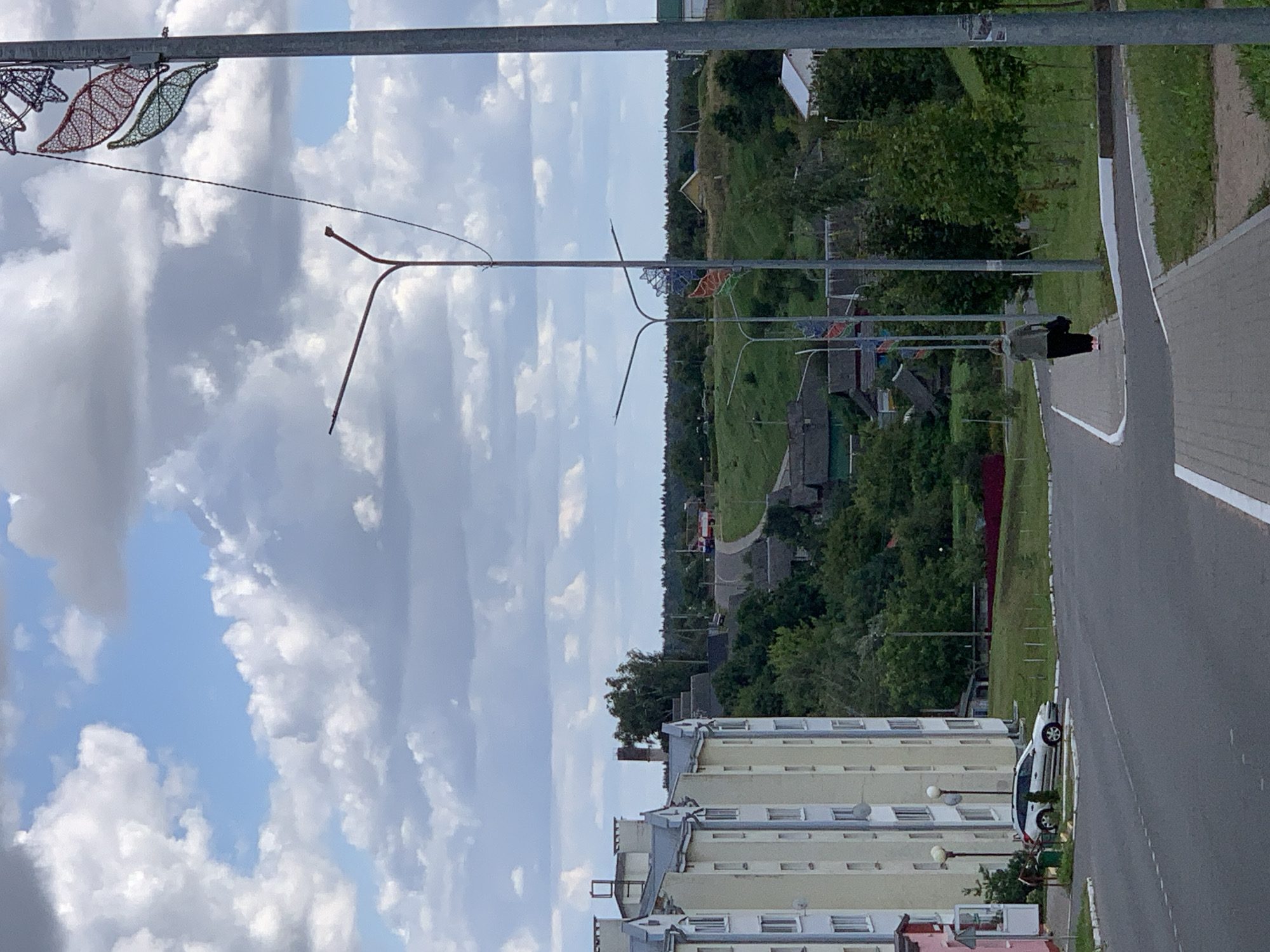 This is a vertical, eye-level, outdoor, daytime photograph taken in the city of Kapyĺ, Belarus. The image captures a residential street scene with several buildings, trees, and a few people.
In the foreground, a paved road with white lane markings curves to the left. A sidewalk runs parallel to the road on the right. A single woman, wearing a green hooded jacket and black pants, walks away from the camera on the sidewalk. She carries a dark bag over her left shoulder and wears pink shoes.
To the left of the road, a multi-story apartment building with a light yellow facade and white trim dominates the left side of the frame. Several windows are visible, and there are balconies on the upper floors. A white car is parked on the grass in front of the building.
In the middle ground, a winding road leads up a hill, past a collection of older, low-rise houses with dark roofs. Green trees and foliage are abundant throughout this area. A tall, narrow brick chimney is visible among the trees in the distance, behind the apartment building.
The background features a distant landscape of rolling green hills covered in trees. A few scattered houses with yellow roofs are visible on the hills. The sky is partly cloudy with large, white cumulus clouds against a light blue sky.
Along the right edge of the image, a tall, gray metal lamppost is visible, adorned with decorative light fixtures resembling leaves and other geometric shapes, illuminated with red, green, and blue lights. Another lamppost with a curved arm is seen in the middle ground, to the left of the decorative one.