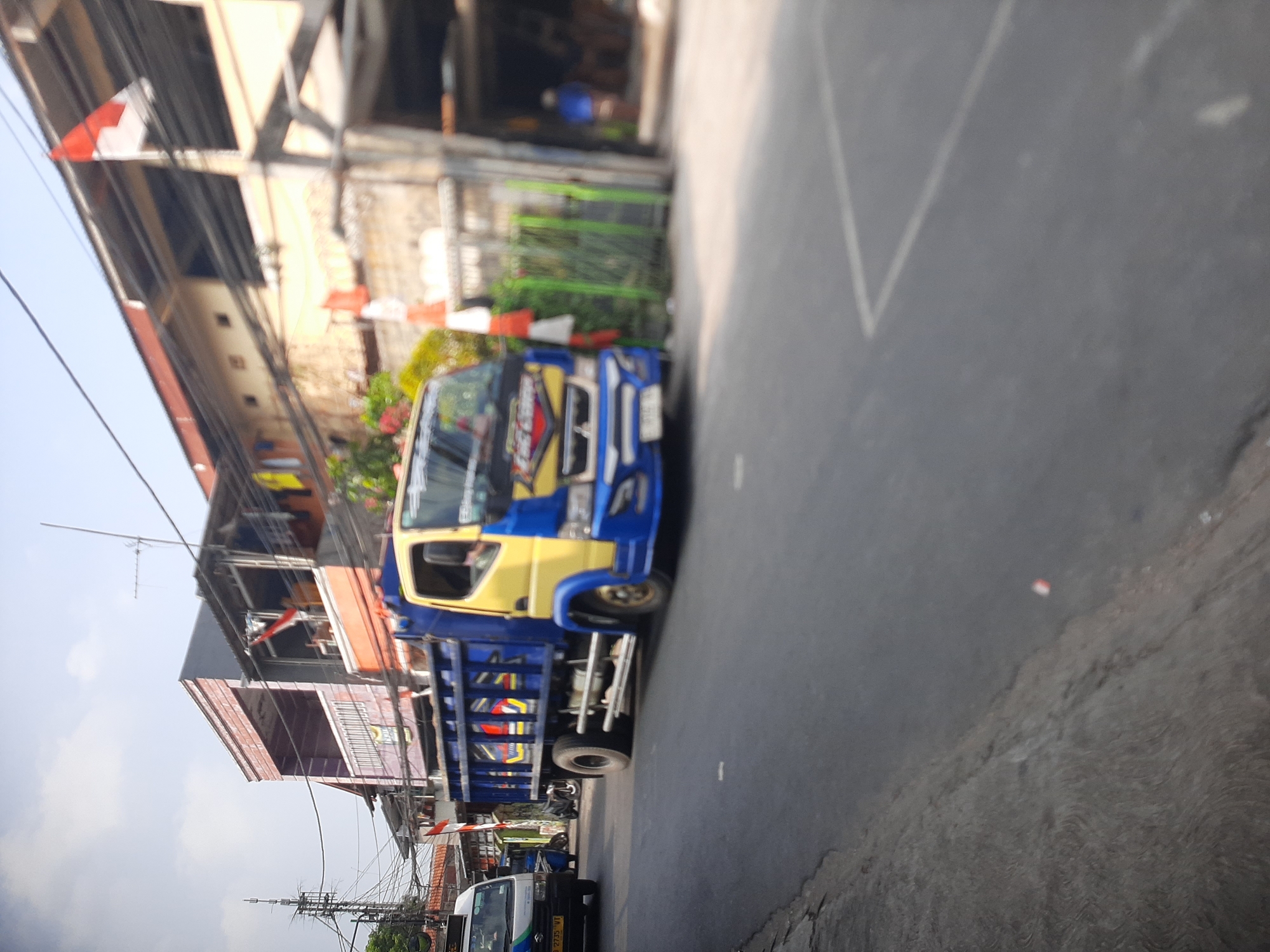 A yellow and blue dump truck is parked on an asphalt road, facing right. The truck has a visible license plate that reads "B 9927 VT". The truck's bed is empty. To the left of the dump truck, a white van with a license plate "B 2735 VT" is also parked. In the background, there are buildings with multiple stories and various signs. An Indonesian flag is visible flying from one of the buildings. The sky is overcast. This scene is likely located in Central Jakarta, Indonesia, given the common vehicle license plate format and the presence of an Indonesian flag.
