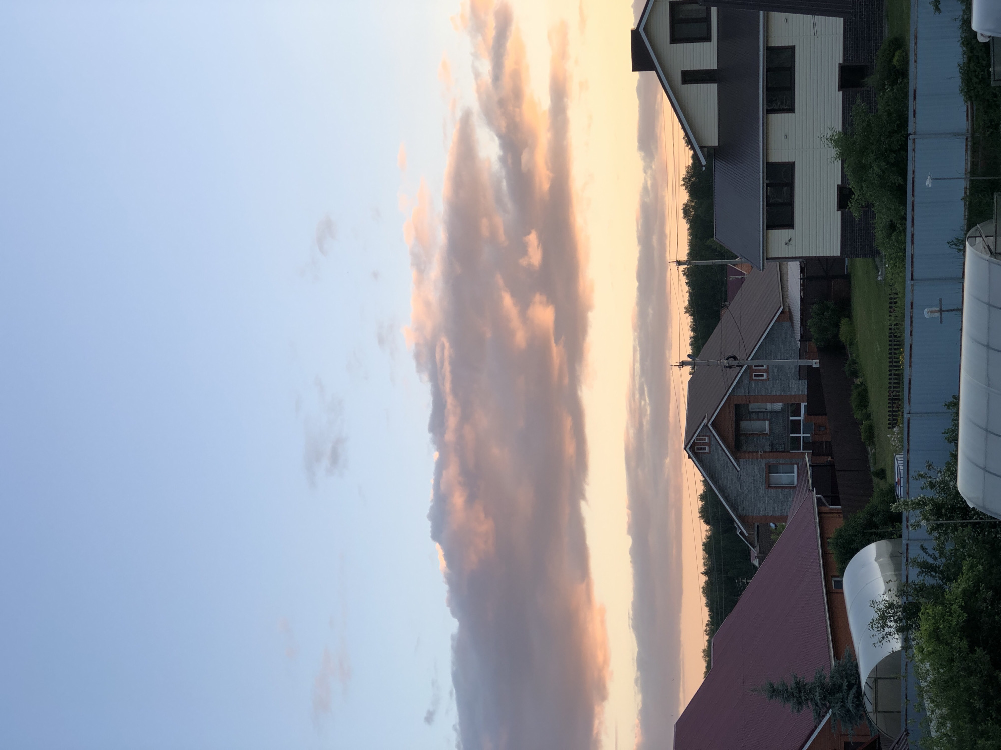 The image depicts a suburban landscape with several houses and clear sky. The sky is a gradient of light blue at the top, transitioning to a soft orange and pink hue near the horizon, indicating either sunrise or sunset. Several large clouds are illuminated by the warm light, creating a picturesque scene.
In the foreground, there are several residential buildings. The house on the far left has a distinctive dark red, sloped roof and appears to be constructed with brick on the lower levels. To its right, a silver, arched greenhouse-like structure is visible.
Further back, a house with a gray stone facade and a brown roof is prominent. This house has multiple windows, some with dark frames, and a balcony. Adjacent to it and slightly to the right is a larger, two-story house with a light-colored siding and a dark roof. This house has a chimney and several rectangular windows.
Behind the houses, a dense line of trees forms a dark green backdrop against the sky. Utility poles with wires are also visible, suggesting the presence of infrastructure. The ground in front of the houses is covered with green grass, and fences separate different properties. The overall impression is of a tranquil residential area. The location is identified as Noviy, Russia.