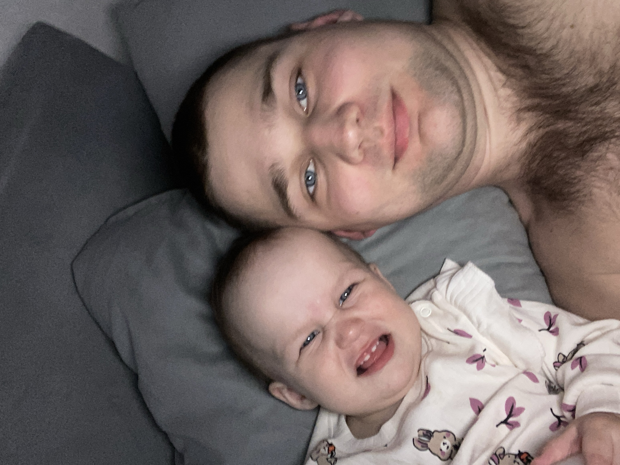 A photo captures a man and a baby lying down on what appears to be a grey pillow. The man is fair-skinned with short dark hair and blue eyes. He has facial stubble and chest hair is visible. The baby is fair-skinned, wearing a white pajama top with a pattern of purple leaves and brown bears. The baby is making a face, scrunching up their nose and showing their teeth. The background is blurred, but suggests an indoor setting. The image was taken in the city of Chaikovsky, Russia.