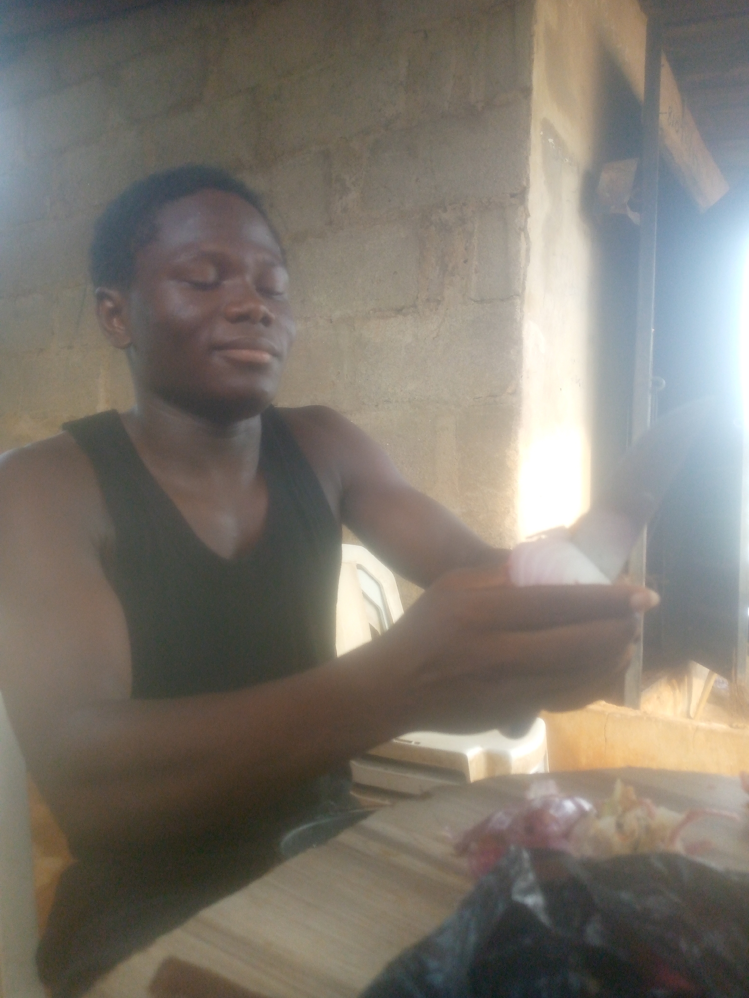 A young person, likely male, with dark skin and short dark hair, is seated and engaged in food preparation. They are wearing a black sleeveless top or tank top. Their eyes are closed, and there's a slight, serene expression on their face, possibly due to the bright light or concentration.
The primary activity is chopping or peeling an onion. The person is holding a white onion in their left hand and a silver knife in their right, positioned as if cutting. On the light-colored wooden cutting board in front of them, there are already chopped pieces of red/purple onion and possibly other pale food items. A crumpled black plastic bag is also on the table, to the right of the chopped ingredients.
The setting appears to be an indoor or semi-enclosed area with rough, light-colored concrete block walls. A strong, bright light source, possibly sunlight from a window or opening, is casting a glow from the right side of the frame, creating overexposed areas and harsh shadows. A dark wooden beam is visible near the top right corner. A light-colored chair is partially visible behind the person.
The image suggests daytime. There is no visible text.
