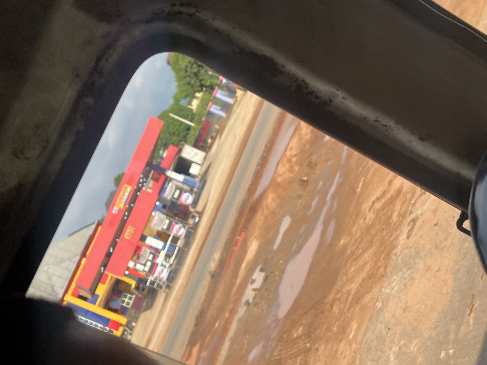 The image captures a scene through a car window, focusing on a colorful building that appears to be a gas station. The building is predominantly red with accents of yellow and blue. The gas pumps and the station's canopy are clearly visible. In the foreground, the ground is muddy with several puddles of water, suggesting recent rain or poor drainage. The overall environment seems to be an outdoor urban or semi-urban setting in Awkuzu, Nigeria, with a road leading to the gas station and some greenery in the background. The perspective indicates the photo was taken from inside a vehicle, looking out.