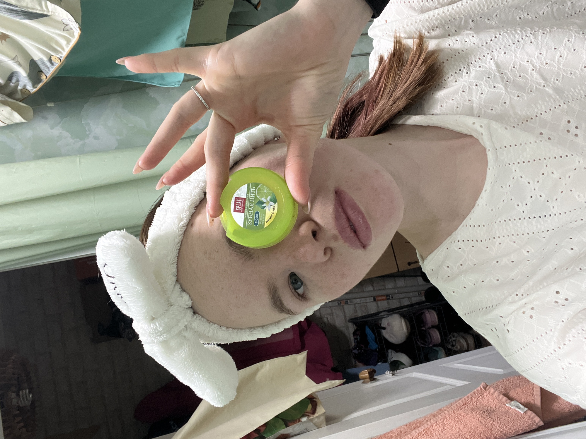 A woman is posing indoors, possibly in Chaikovsky, Russia given the Russian text on the product she is holding. She is wearing a white, eyelet-patterned top and a white headband with bunny ears. Her hair is reddish-brown. She is holding a round, green container with Russian text and an image of a lime and flower in front of her eye. Her hand is outstretched above the container with a ring visible on one finger. Background elements suggest a bed or couch with pillows and blankets, as well as a door and storage rack with various items. The woman appears to be the primary subject of the image, possibly taking a selfie.
