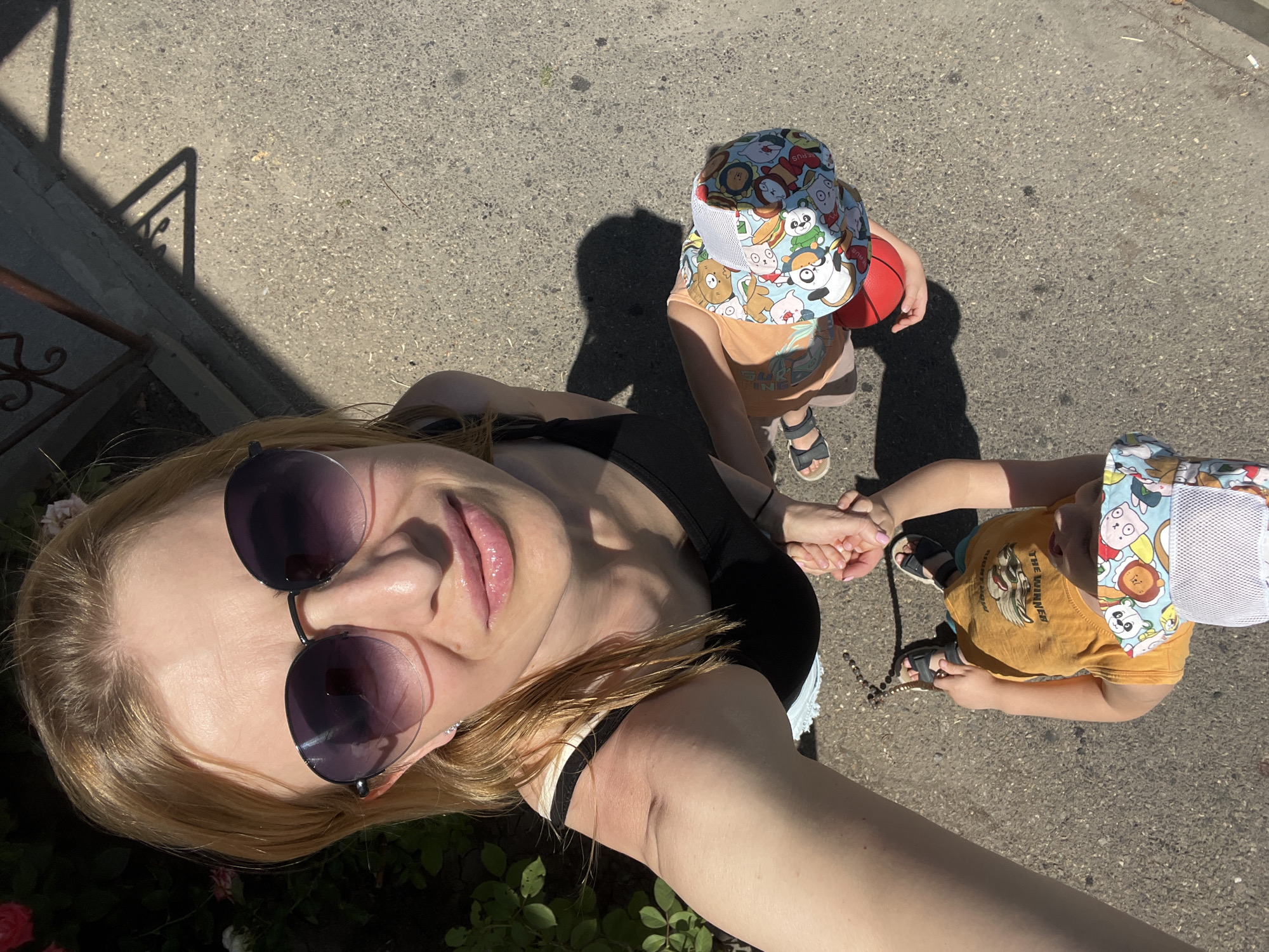 A woman with blonde hair and sunglasses takes a selfie outdoors on a paved surface, likely a sidewalk or street. She is wearing a black tank top and white shorts. In front of her, two children are visible. One child, wearing a colorful hat with cartoon characters and an orange shirt, is being held by the woman. The other child, wearing an orange t-shirt with a lion graphic and sandals, is standing and holding a small red ball. The background includes greenery, possibly rose bushes, and a glimpse of a metal railing and shadow from a structure, suggesting an urban or suburban environment. The image appears to be taken on a sunny day. No specific landmarks or identifying features definitively place the scene in Bataysk, Russia.