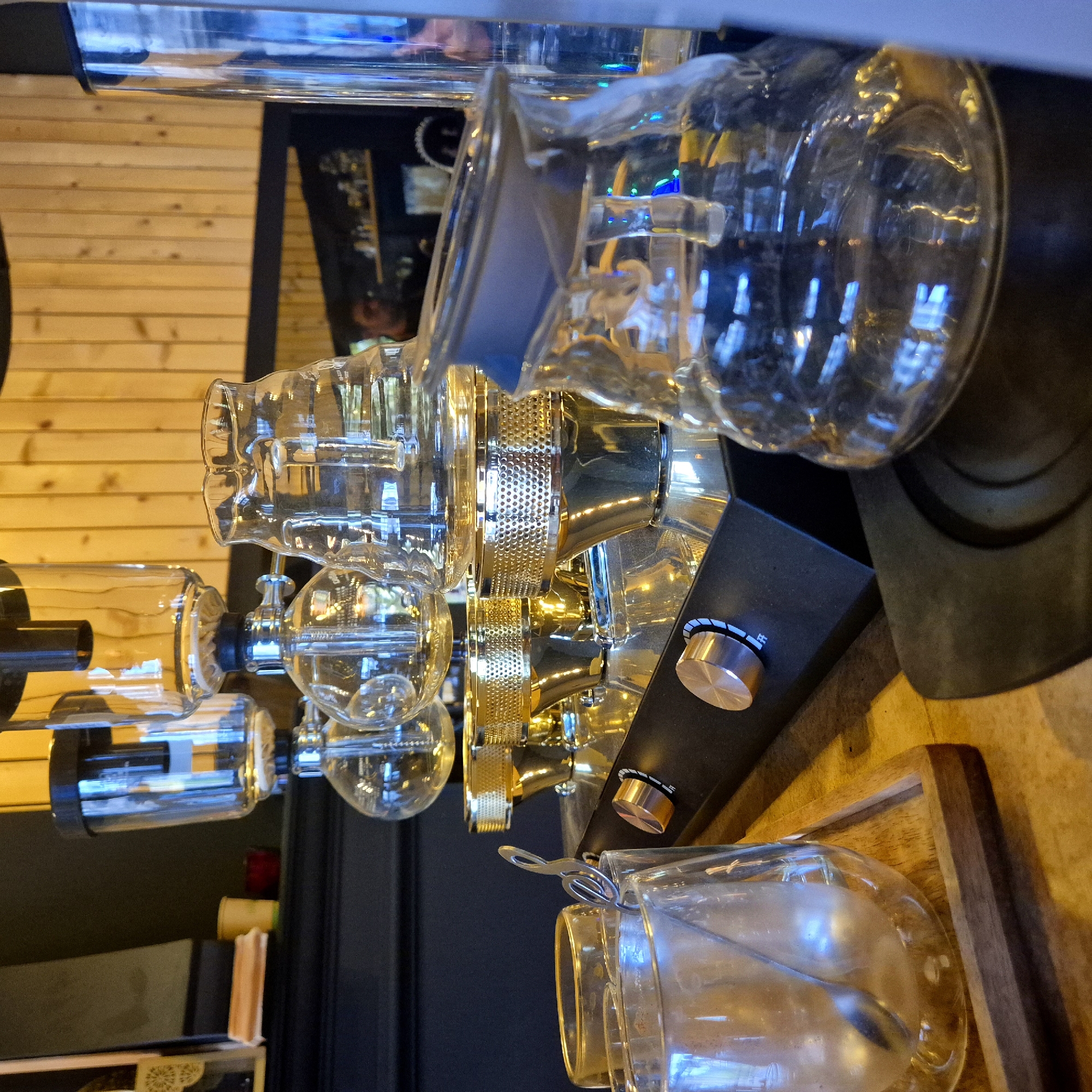 The image shows a close-up of a high-end cold brew coffee brewing system. The system consists of several glass bulbs and tubes connected by metal components. There are two prominent silver knobs on a dark base. In the foreground, there is a glass jar containing liquid with a metal spoon or stirrer inside. The background includes wooden paneling and a dark area with some reflection, possibly indicating a window or mirror. The location is likely a coffee shop in the city of Strasbourg, France, due to the system's design and prevalence in such establishments.