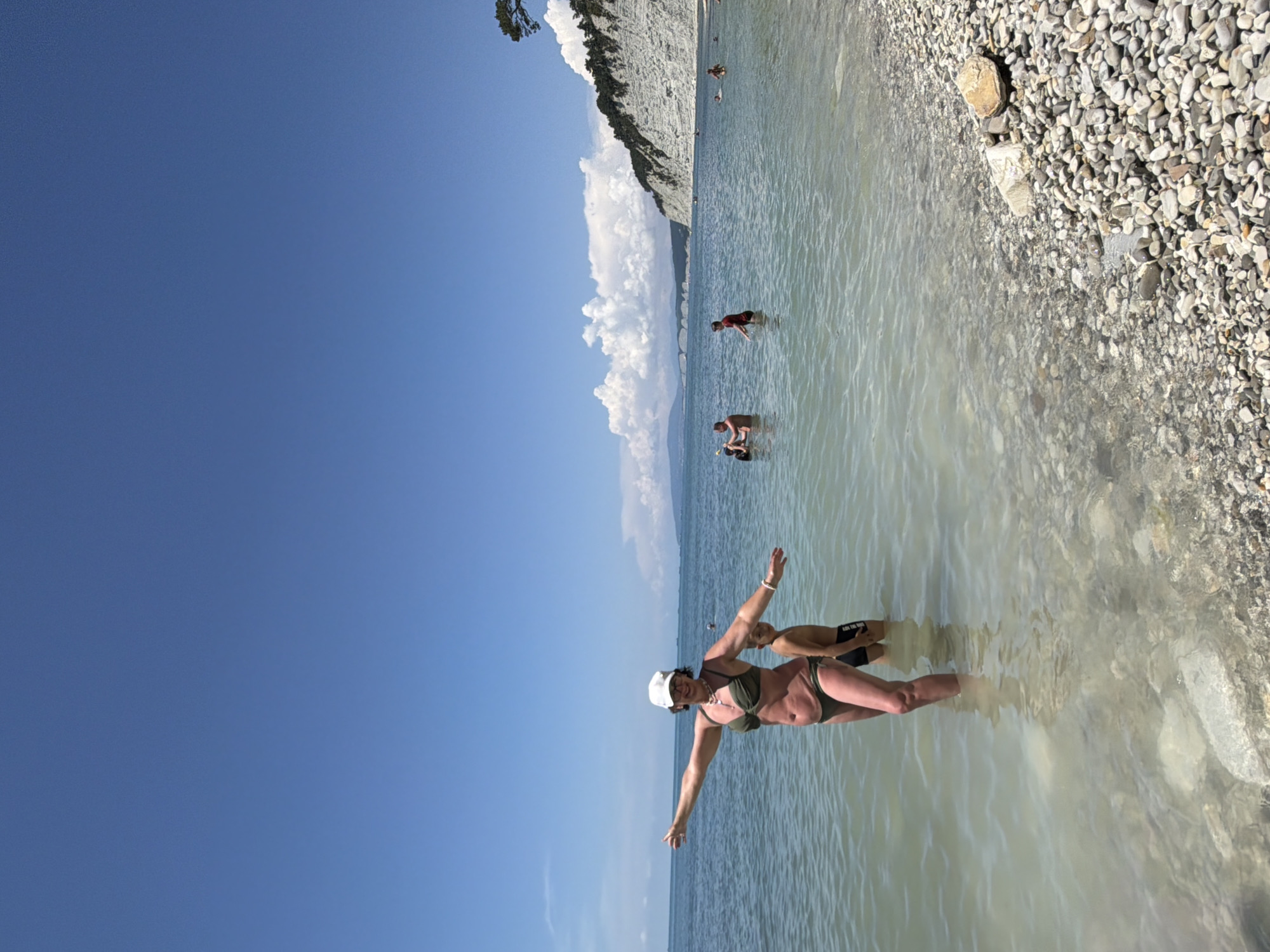 A woman in a green bikini and a white baseball cap stands in shallow, clear ocean water with her arms outstretched. A boy in dark swim trunks stands behind her. The beach is covered in smooth, grey pebbles. In the background, other people are visible in the water, and a large, white cliff face covered in trees rises from the coastline. The sky is a clear, bright blue with a few scattered white clouds. The scene appears to be the coast of Divnomorskoe, Russia.