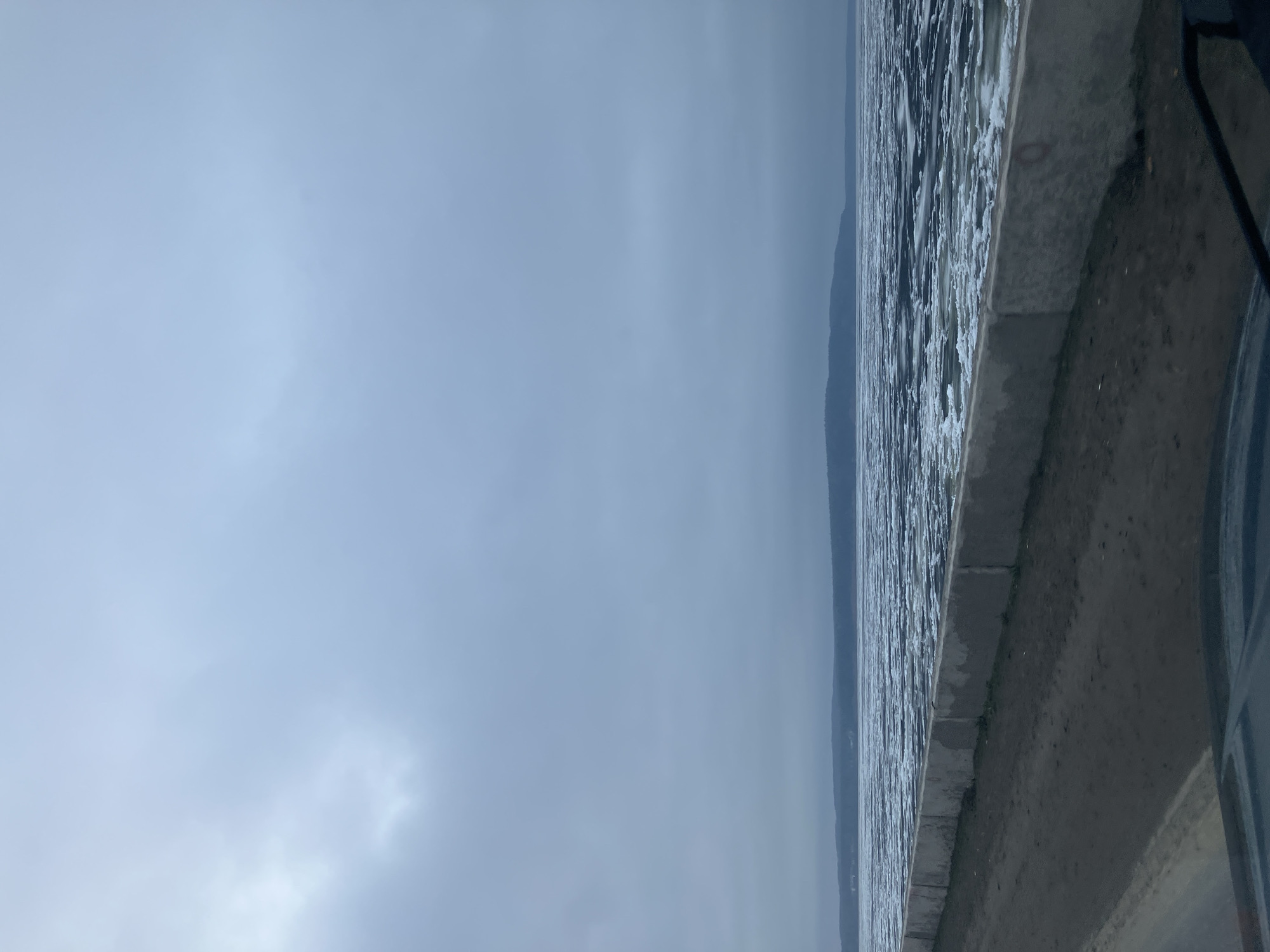 The image presents a wide, expansive landscape view of a large body of water, likely a lake or river, under an overcast sky. In the foreground, a grey concrete embankment or curb runs horizontally along the bottom of the frame, bordering a paved or dirt pathway. A dark, indistinct object, possibly part of a vehicle or structure, is partially visible on the far right, suggesting the photographer's immediate surroundings.
The water's surface, located to the left of the embankment, displays numerous ripples and small waves, reflecting the soft, muted light of the sky. It extends towards a hazy horizon in the midground, where a faint, dark landmass—appearing as distant hills or mountains—is subtly silhouetted against the lighter sky.
The sky occupies the upper portion of the frame, characterized by uniform light grey, dense clouds. There are no patches of blue sky or direct sunlight visible, indicating heavily overcast weather conditions. Some subtle variations in cloud density can be observed. The overall color palette is dominated by cool tones of grey and blue, contributing to a serene and somewhat somber atmosphere.
No people, animals, or specific activities are depicted in the scene, lending a tranquil and solitary feel. It appears to be daytime under cool or cold weather. The presence of the concrete embankment suggests a developed waterfront area, consistent with a setting in Chaikovsky, Russia. No visible text is present in the image.