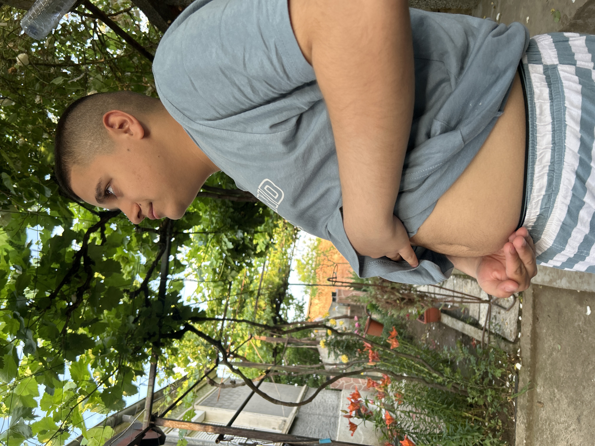 A young person, possibly male, is depicted from the waist up in an outdoor setting, viewed from a slightly elevated angle. They have short, dark hair styled in a fade haircut and are wearing a light blue-grey short-sleeved t-shirt and blue and white striped shorts. Their gaze is directed downwards and slightly to their left, with a neutral or pensive expression.
The person's right hand is holding the bottom hem of their t-shirt, pulling it up slightly to reveal their lower abdomen. Their left arm is bent, with the hand also near their midsection, possibly adjusting clothing or resting. A faint, white graphic or logo is present on the front of the t-shirt, but it is not clear enough to be deciphered.
The background is filled with lush green foliage, likely from a climbing vine, creating a natural canopy overhead. Through the leaves, patches of bright blue sky are visible, indicating a clear, sunny day. A clear plastic water bottle is partially visible, hanging upside down among the leaves in the upper left. Below the foliage, parts of a building with a light-colored roof and stone facade can be seen, suggesting a residential or garden environment. Various potted plants, including vibrant orange lilies, are arranged on the ground, which appears to be a mix of concrete and soil. A metal trellis or support structure for the vines is also discernible. The bright, natural light suggests it is daytime, possibly morning or early afternoon.