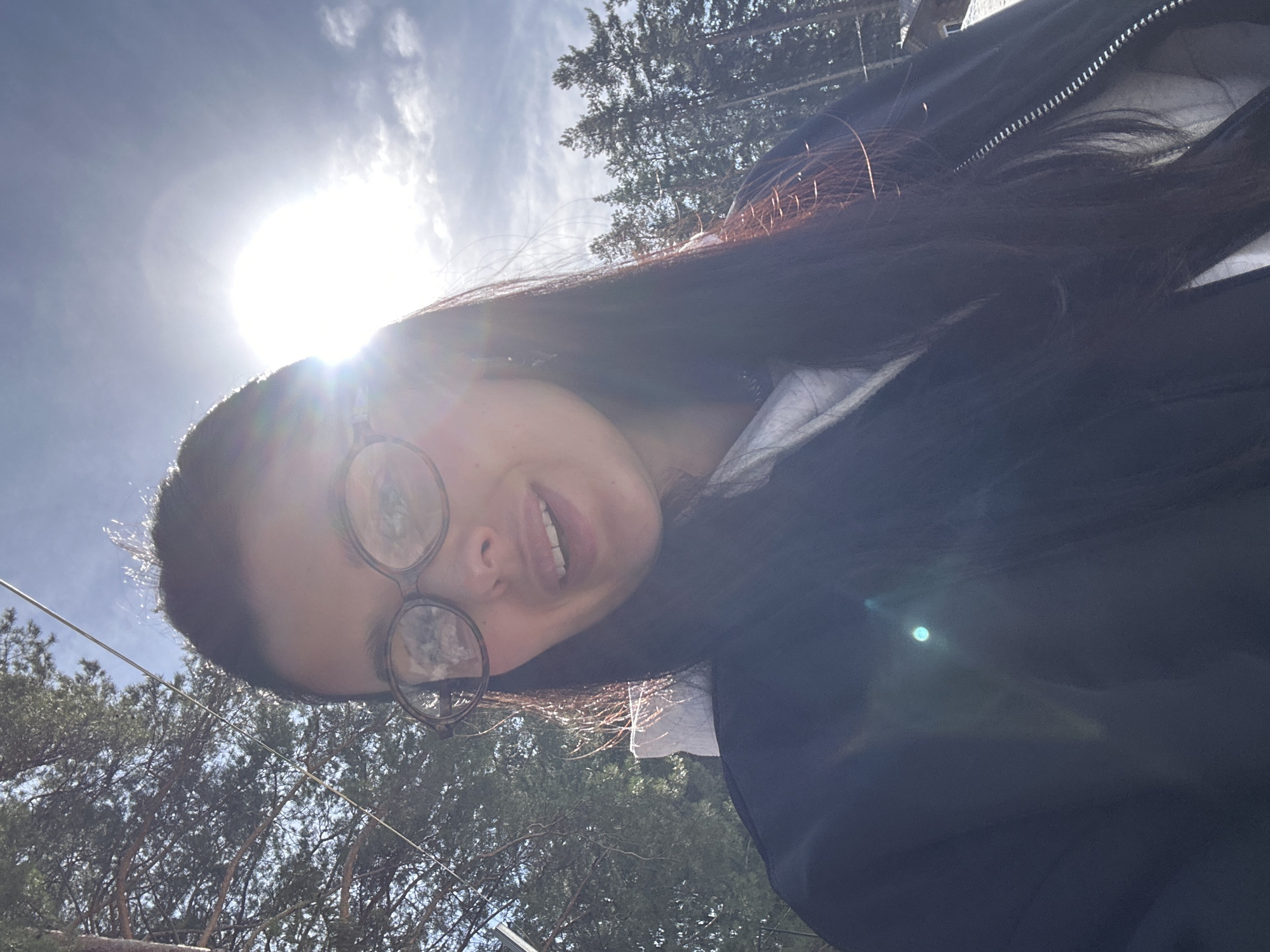 A low-angle shot of a person wearing glasses against a sunny sky, possibly outdoors in the Прикамский, Russia. A black jacket covers a white hoodie. Trees are visible in the background. The person is smiling, and the sun is causing lens flare.