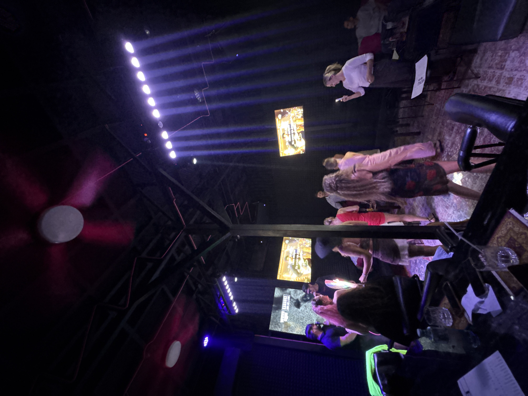 The image depicts a lively indoor scene, likely a karaoke bar or entertainment venue in Baan Bang Rak, Thailand. The ambiance is illuminated by a combination of stage lights, creating dramatic beams of purple and pink light, and overhead lighting.
In the foreground, a dark table displays several glasses, a bottle of water, and some papers, suggesting a seating area or bar counter. The middle ground features a group of people, some dancing and socializing. A woman with long blonde hair is prominently visible, wearing a dark floral dress. Another woman in a pink dress is also noticeable.
Behind the people, two large screens are mounted on the wall, displaying karaoke lyrics or visuals, further confirming the nature of the establishment. The ceiling is a dark, industrial design with exposed beams and electrical wiring. A large fan with pink blades is suspended from the ceiling, contributing to the overall atmosphere.
In the background, more patrons are seated and standing, enjoying the entertainment. A woman in a white shirt and long skirt is standing on the right, holding a phone, possibly recording or taking photos. The overall impression is one of a social gathering, filled with music and entertainment in a dimly lit, modern venue.