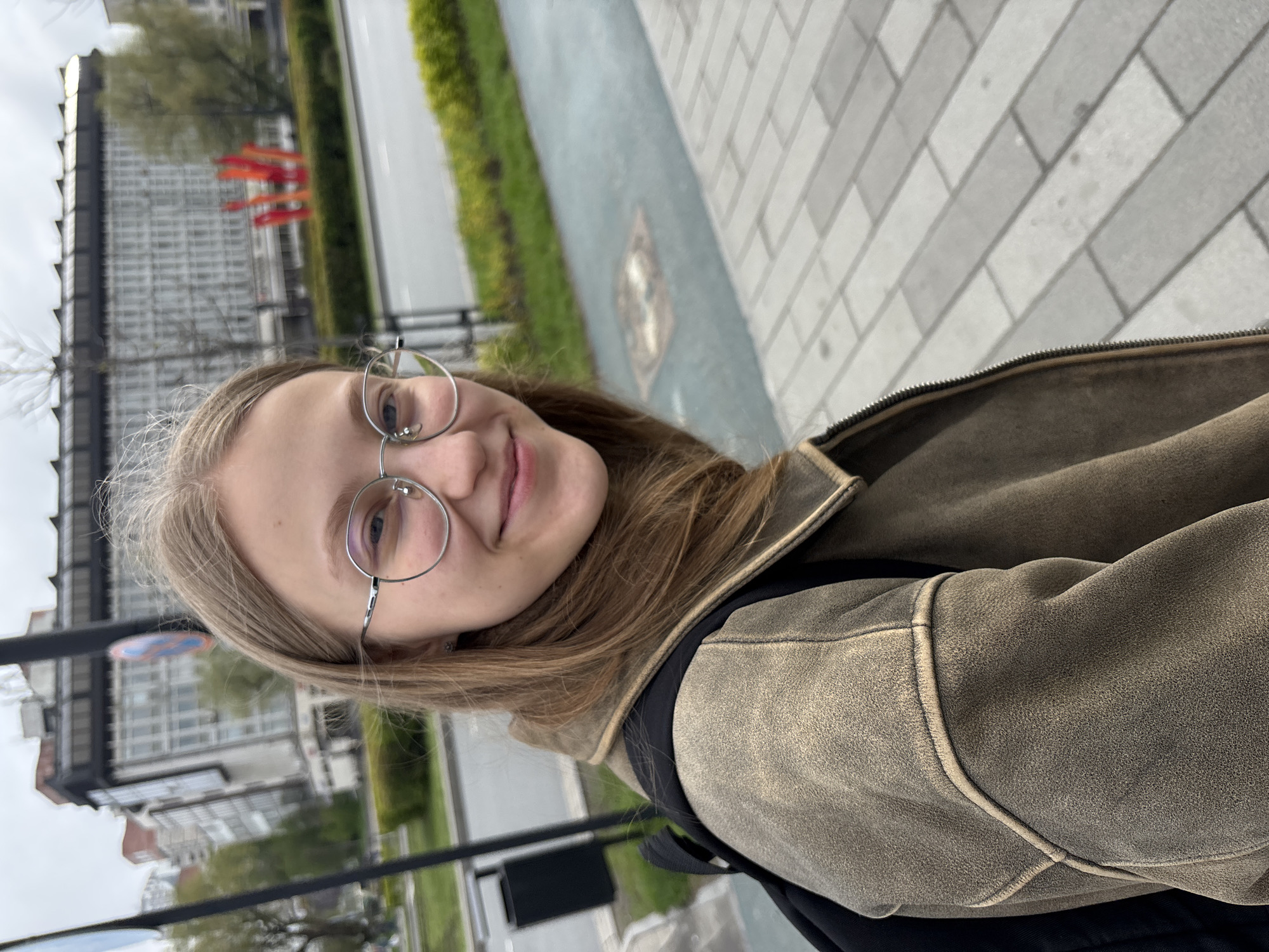 A young woman with light brown hair and round glasses smiles at the camera. She is wearing a brown jacket with black accents and a black backpack. The photo appears to be taken on a city sidewalk. In the background, buildings and greenery are visible, along with what may be red flags or banners. Given the location of Perm, Russia, the buildings in the background may be part of the city's architectural landscape. The time of day is likely daytime.