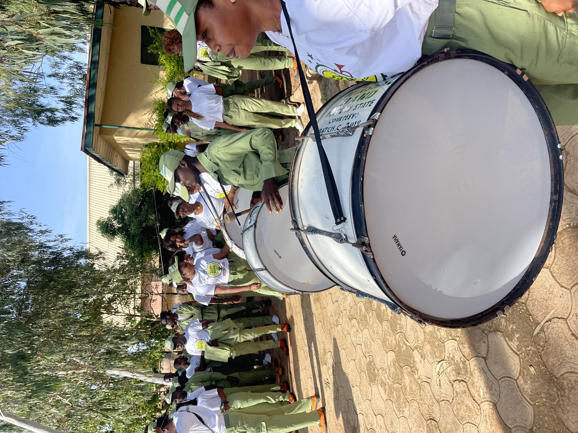 A group of young adults, likely participating in a community service program given their matching green uniforms and white t-shirts, are gathered outdoors. The uniforms consist of green caps, shirts, and trousers, with some individuals wearing white t-shirts with an emblem that includes the text "NYSC BAND." They are holding and interacting with large bass drums, suggesting they are part of a band or musical ensemble. The environment appears to be a paved area with a patterned brick surface, possibly a courtyard or walkway, surrounded by trees and a building with a light-colored facade and a dark-tiled roof. The sky is clear and blue, indicating daytime. The overall scene suggests an organized event or practice session in an outdoor urban setting. The image is likely taken in Jos, Nigeria, a city known for its cultural diversity and community programs.
