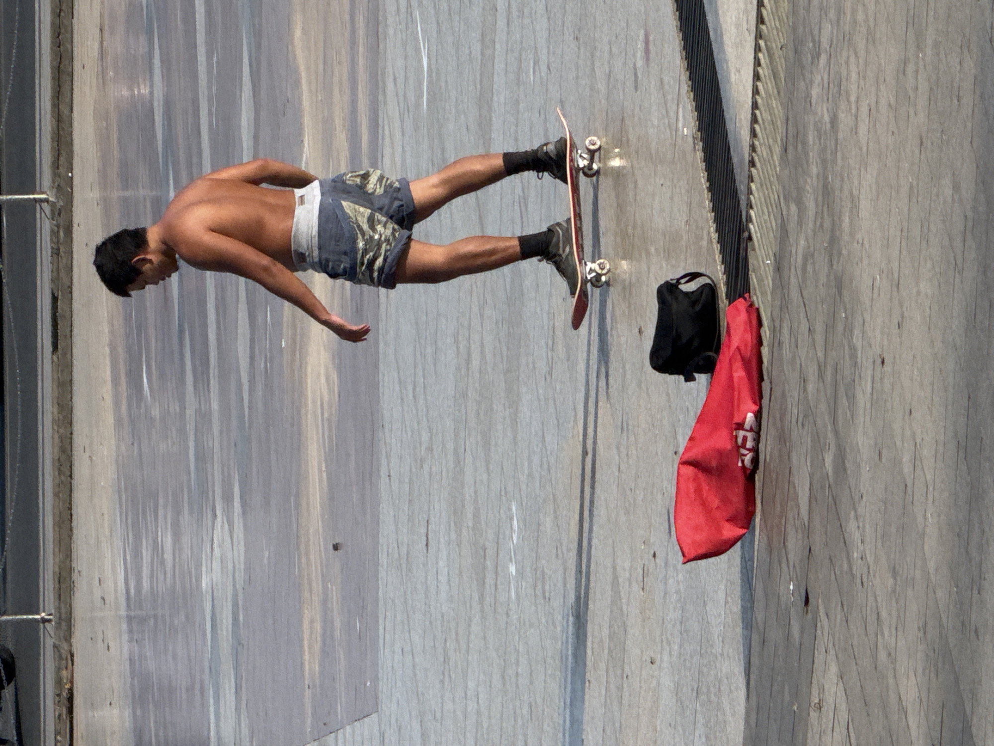A shirtless young man with dark hair rides a skateboard on a smooth, light-colored paved surface. He is wearing camouflage patterned shorts and black socks and shoes. In the foreground, to the left of the skateboarder, lies a crumpled red fabric object and a black bag. The environment appears to be an outdoor public space, possibly a plaza or park, with a grate visible to the right. The lighting suggests it is daytime with sunlight casting shadows. The location is the Xuan Huong Lake area in Da Lat, Vietnam, known for its wide, flat pavements suitable for activities like skateboarding.
