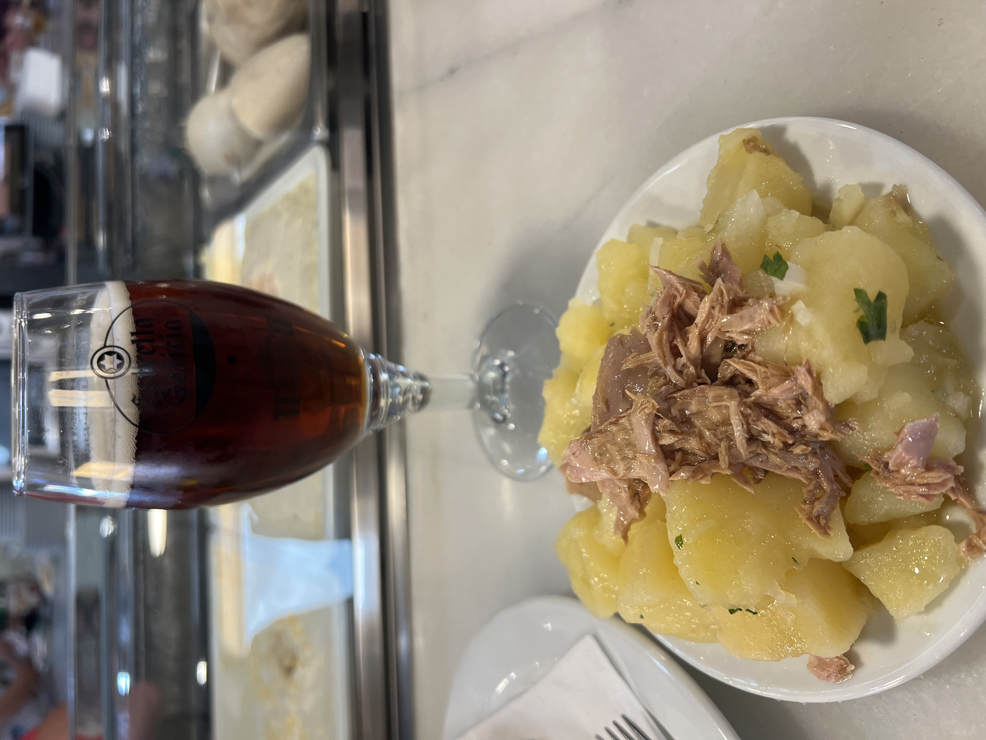 The image shows a close-up view of a plate of potato salad topped with tuna and a glass of dark beer. The beer glass has a logo that reads "Estrella Galicia". The food is presented on a white plate, and the background appears to be a restaurant or bar setting, with other plates of food visible behind the beer glass. The overall lighting suggests an indoor environment during the day. The scene is likely a tapas bar in Sanlúcar de Barrameda, Spain, a region known for its seafood and sherry, where Estrella Galicia beer is also popular.