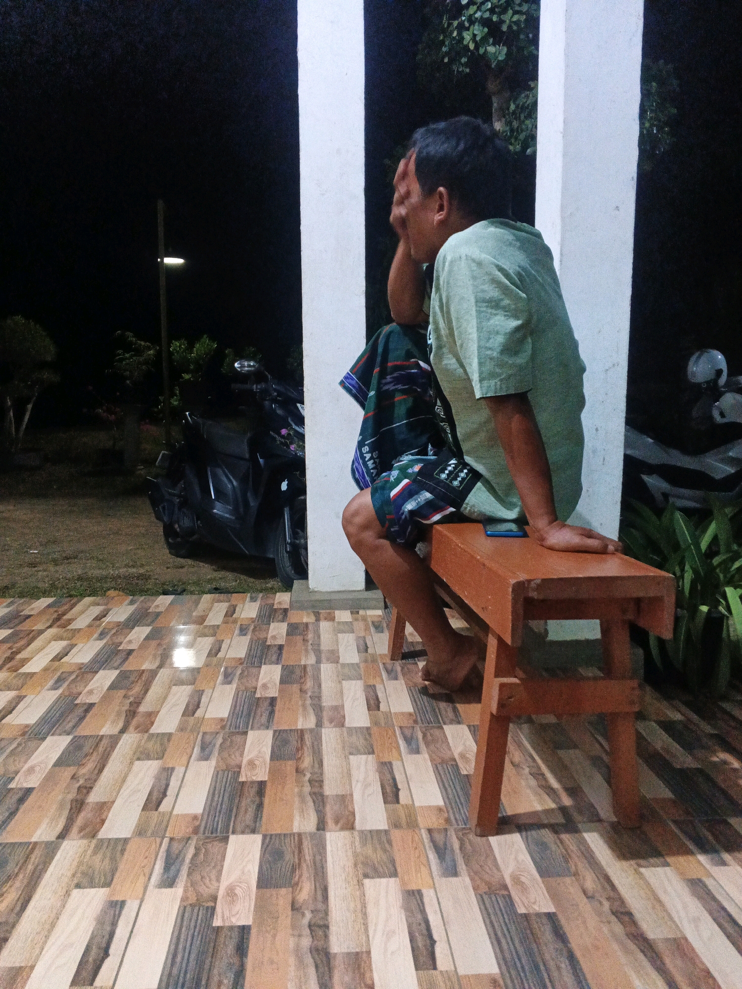 The image depicts a nighttime scene in Desa Gajah Mati, Indonesia. A man is seated on a wooden bench, facing away from the camera and towards the right. He is wearing a light green t-shirt and patterned shorts, with a dark patterned cloth draped over his left shoulder. His right hand is raised to his forehead, obscuring his eyes. The bench is positioned on a tiled floor with a wood-grain pattern. Behind the man, a dark motorcycle is parked on a grassy area. In the background, there are trees and a lamppost emitting light. Two white pillars are visible on either side of the man and bench. The overall lighting suggests it is after dark.