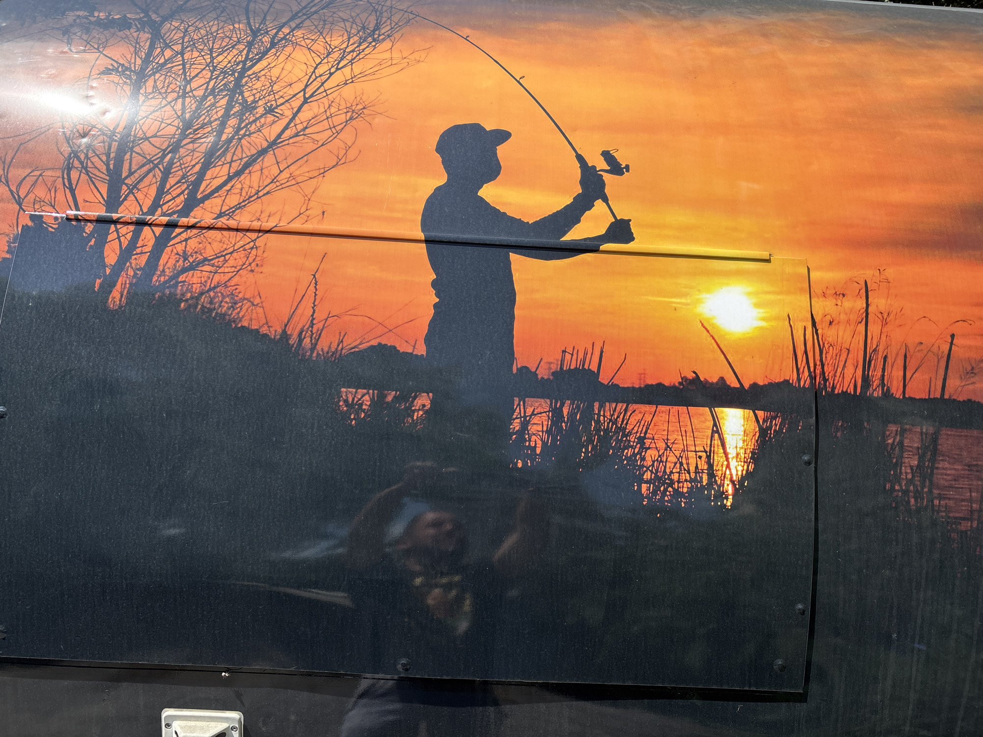 A medium shot shows an image of a fisherman cast against a vivid sunset backdrop affixed to a dark surface. The silhouette of the fisherman, rod raised, is prominent. The setting sun is reflected on the water, with cattails dotting the shoreline. A leafless tree stands on a bank to the left of the fisherman. The dark, reflective surface below the image hints at a possible location on a vehicle or trailer. The bottom portion of the image is marred by a reflection, obscuring further details of the surface. There are no discernable elements indicating the location of Plovdiv, Bulgaria.