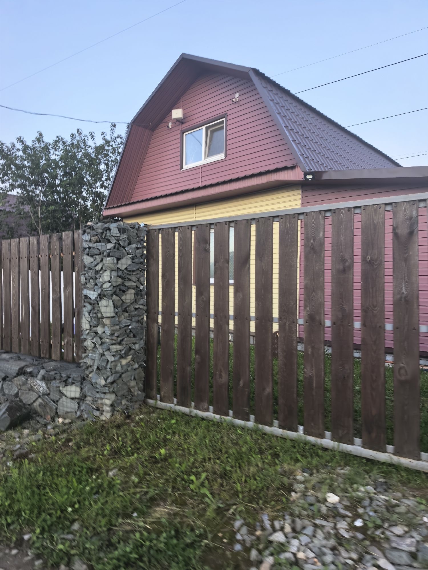 The image shows a house with a dark brown, sloped roof and a red and yellow exterior. A wooden fence with vertical planks runs along the foreground, with a gabion pillar made of stones and wire mesh supporting one end. There is green grass and loose stones in front of the fence, and a clear blue sky above. The house has a single window visible on the upper floor, and two outdoor lights are mounted on the exterior wall near the roofline. Trees are visible behind the fence on the left side of the image. The overall scene suggests a residential property, possibly in a rural or suburban setting.