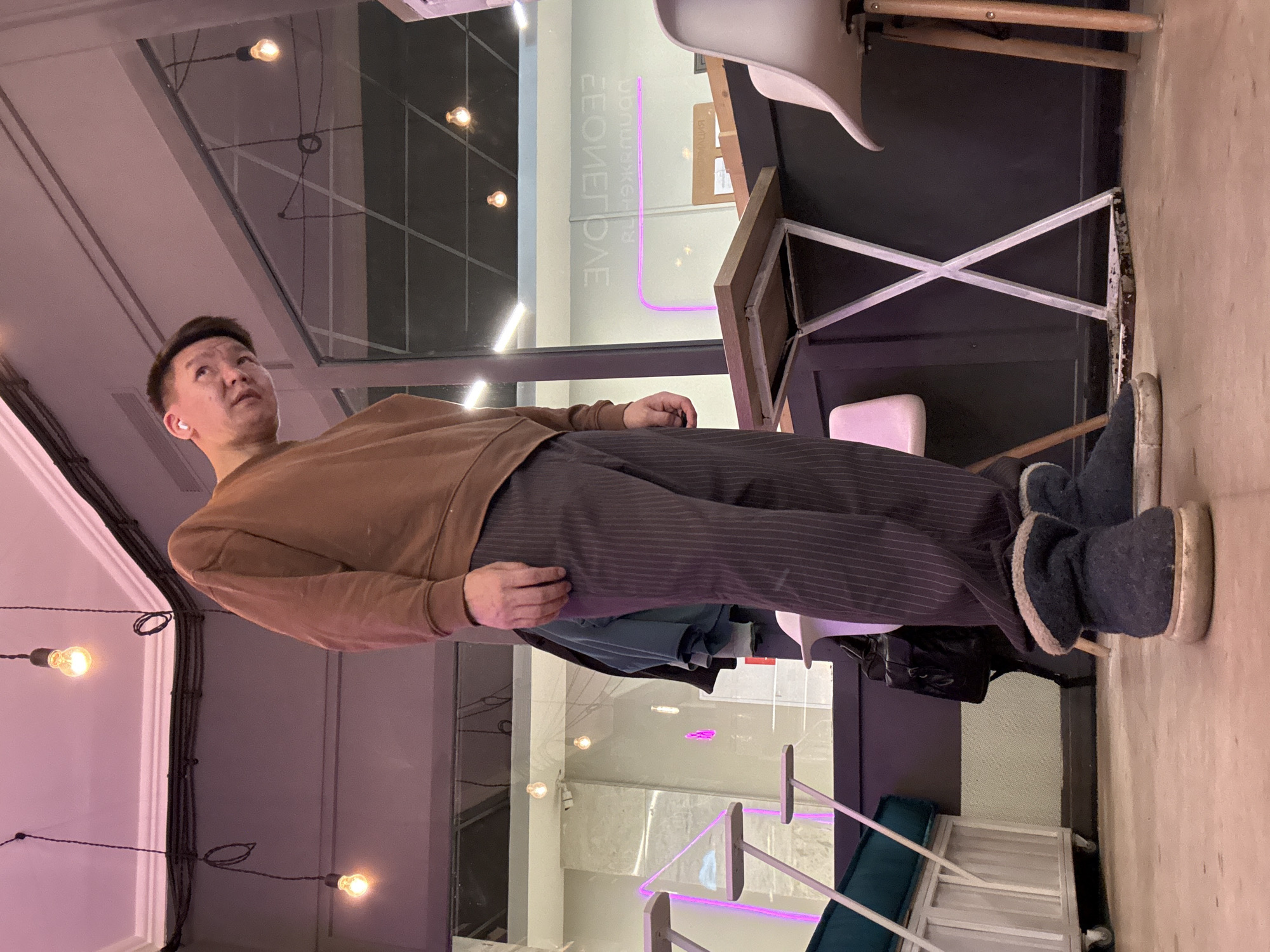 A man stands in an indoor space, likely a clothing store, in Moscow, Russia. He wears a brown sweater, striped pants, and dark-colored boots. He looks upwards. The interior features modern decor, including a white chair with wooden legs, a clothing rack, and decorative lighting. A sign with reversed lettering is visible through a glass partition.