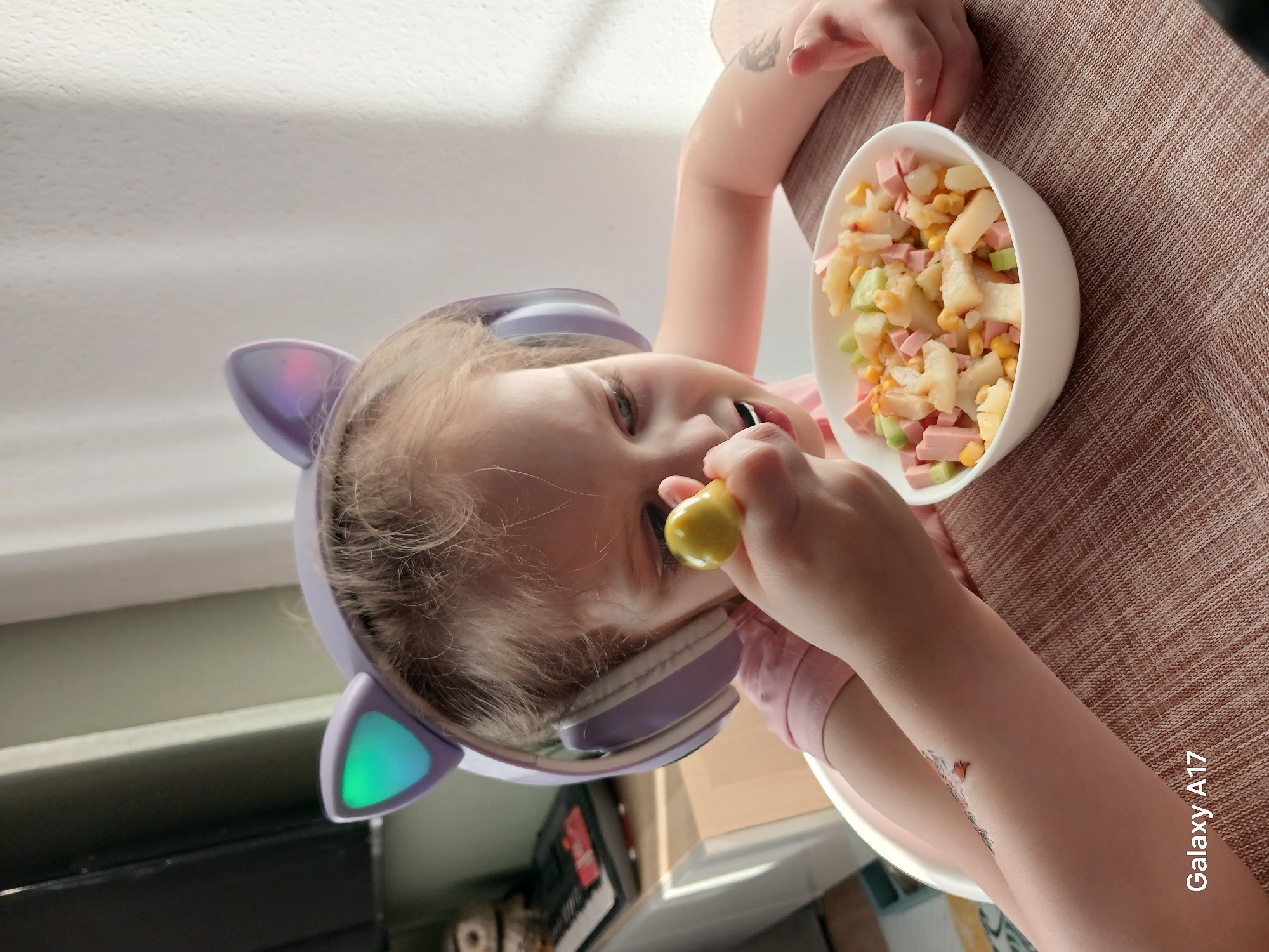 A young child, positioned on a brown textured surface, wears purple cat-ear headphones with illuminated green lights. The child is eating from a white bowl containing a salad of potatoes, ham, corn, and cucumbers. The child holds a yellow object, possibly a toy or piece of food, near their mouth. The surroundings suggest an indoor setting, possibly a home. The location of the image is not discernible from the provided information.