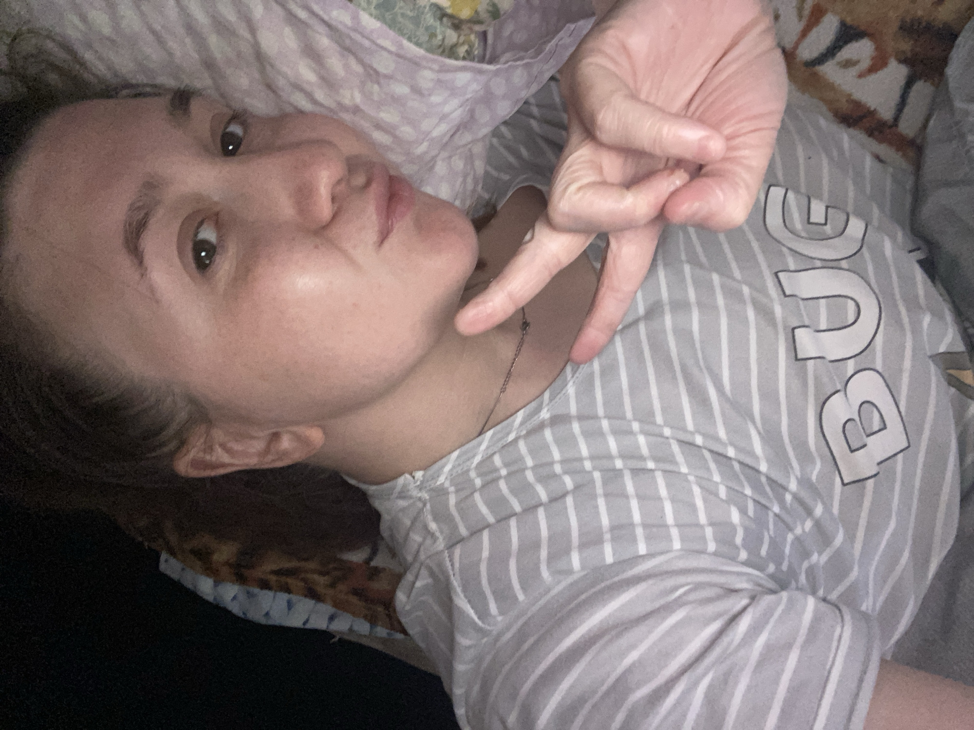 A person is lying down, appearing to be in a bed. The person is facing the camera with their head tilted, and they are making a hand gesture with their right hand. The person is wearing a grey and white striped shirt with the letter "B" visible on it. The bed has different patterned linens. The location is Chaikovsky, Russia.