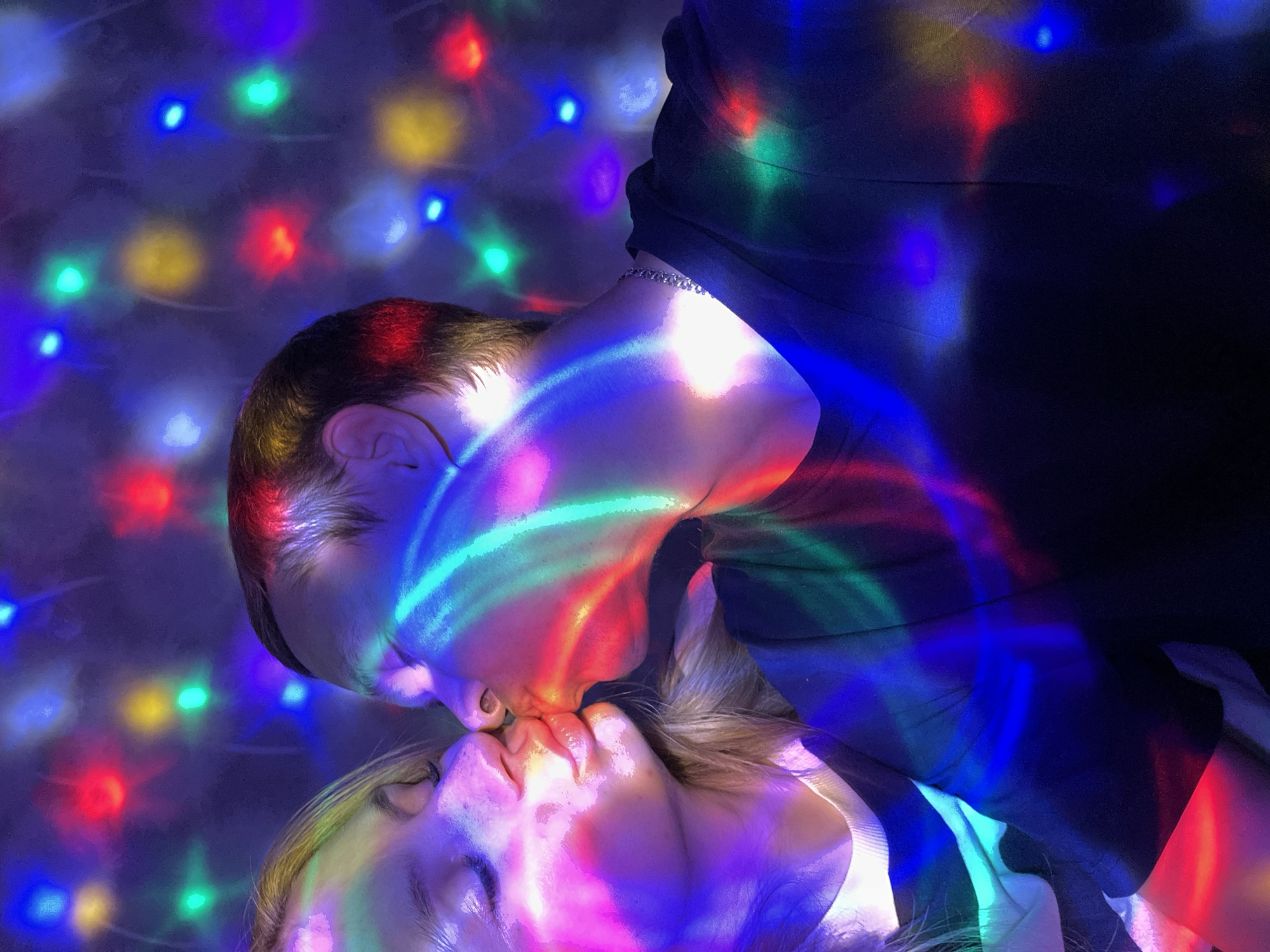 A couple is kissing, illuminated by bright, colorful disco lights. The man is wearing a dark shirt and a silver chain around his neck. The woman has her eyes closed. The background is a blur of colored lights. There is no indication that this image was taken in Chaikovsky, Russia.
