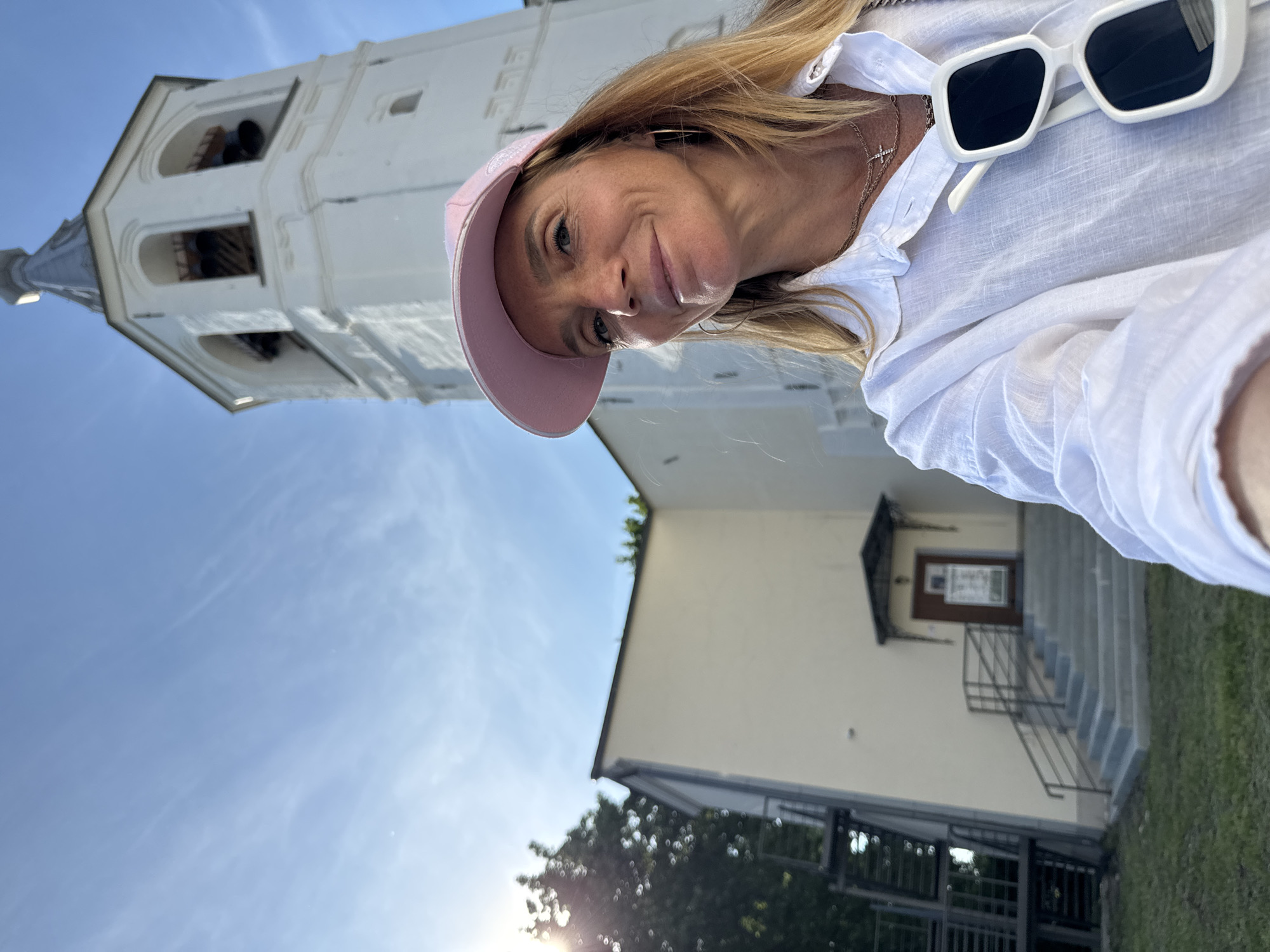 A woman in a white shirt and pink baseball cap takes a selfie in front of a white bell tower with bells visible inside. The bell tower is part of a historical complex in Veliky Novgorod, Russia, likely the Kremlin. The sky is blue with wispy clouds, and sunlight glints off a tree on the left side of the frame. The woman is wearing sunglasses perched on her shirt and a cross necklace. Stairs lead up to a brown door on a building to the left of the bell tower.