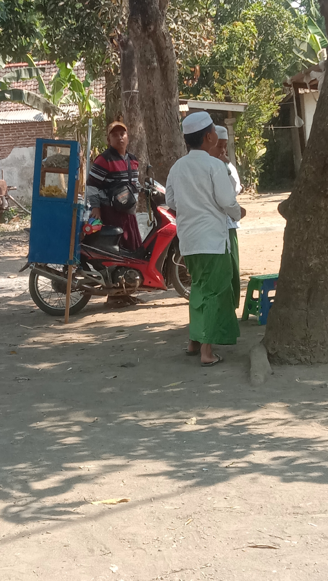 In Ngempit, Indonesia, an outdoor scene unfolds with three individuals and a red motorcycle. A man wearing a striped shirt and a cap stands near a blue cart, which appears to be a food vendor's stall, displaying food items. To his right, two other men, both clad in white shirts and green sarongs, engage in conversation. One of the men is wearing a white cap. The motorcycle, parked beside the vendor's cart, is a prominent feature in the scene. The ground is covered in dirt and debris, and shadows from trees are cast across the area. The background reveals a building with a tiled roof and lush green foliage, suggesting a rural or suburban setting.