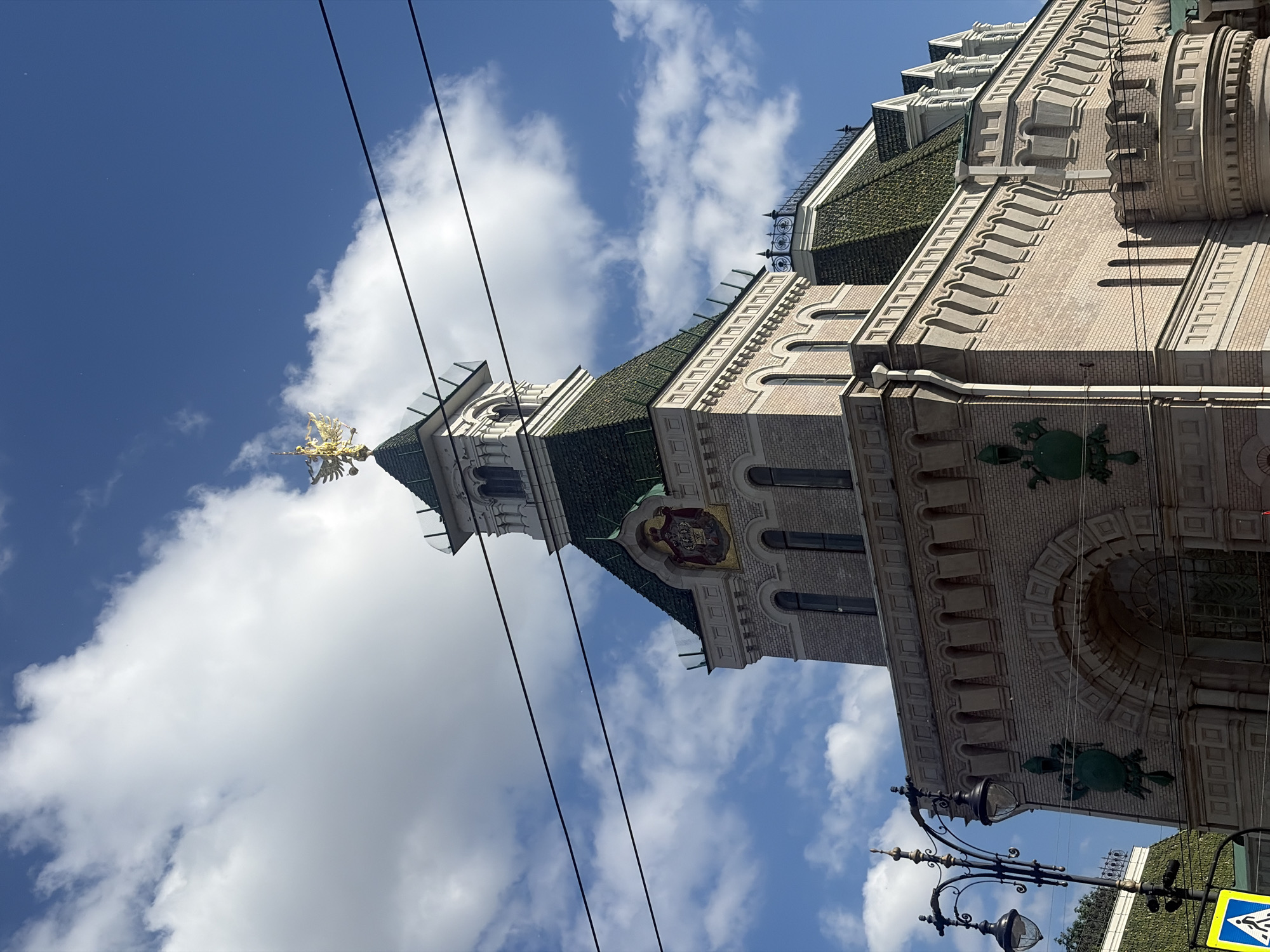 This is a photograph of the Singer House, also known as the Dom Knigi (House of Books), located on Nevsky Prospekt in Saint Petersburg, Russia. The image is taken from a low angle, looking up at the building against a bright blue sky with white, fluffy clouds. The Singer House is an iconic Art Nouveau building, recognizable by its glass dome topped with a golden eagle.
The architecture features a mansard roof covered in green tiles, decorative brickwork, and numerous arched windows. A prominent golden double-headed eagle, the symbol of the Russian Federation, sits atop the spire. Decorative green sculptural elements, possibly stylized animal motifs, are visible on the facade. In the foreground, a black lamppost with two ornate lamps stands to the left, and a yellow pedestrian crossing sign is partially visible at the bottom left. Overhead, dark power lines crisscross the frame. The overall impression is of a sunny day with dramatic architecture.