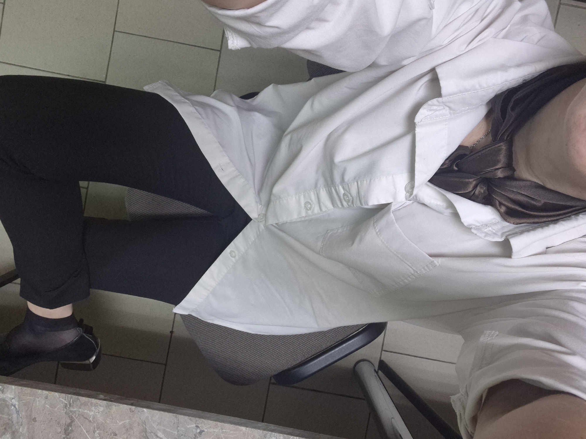 The image shows a close-up, overhead view of a person wearing a white collared shirt, a dark brown scarf tied at the neck, and black leggings. The person is seated on a chair with a textured grey seat and black armrests, positioned near a desk with a marble-like surface. The background consists of light-colored tiled flooring. The setting is indoors, likely an office or study, and there are no visible landmarks or indications of the city of Chaikovsky, Russia.