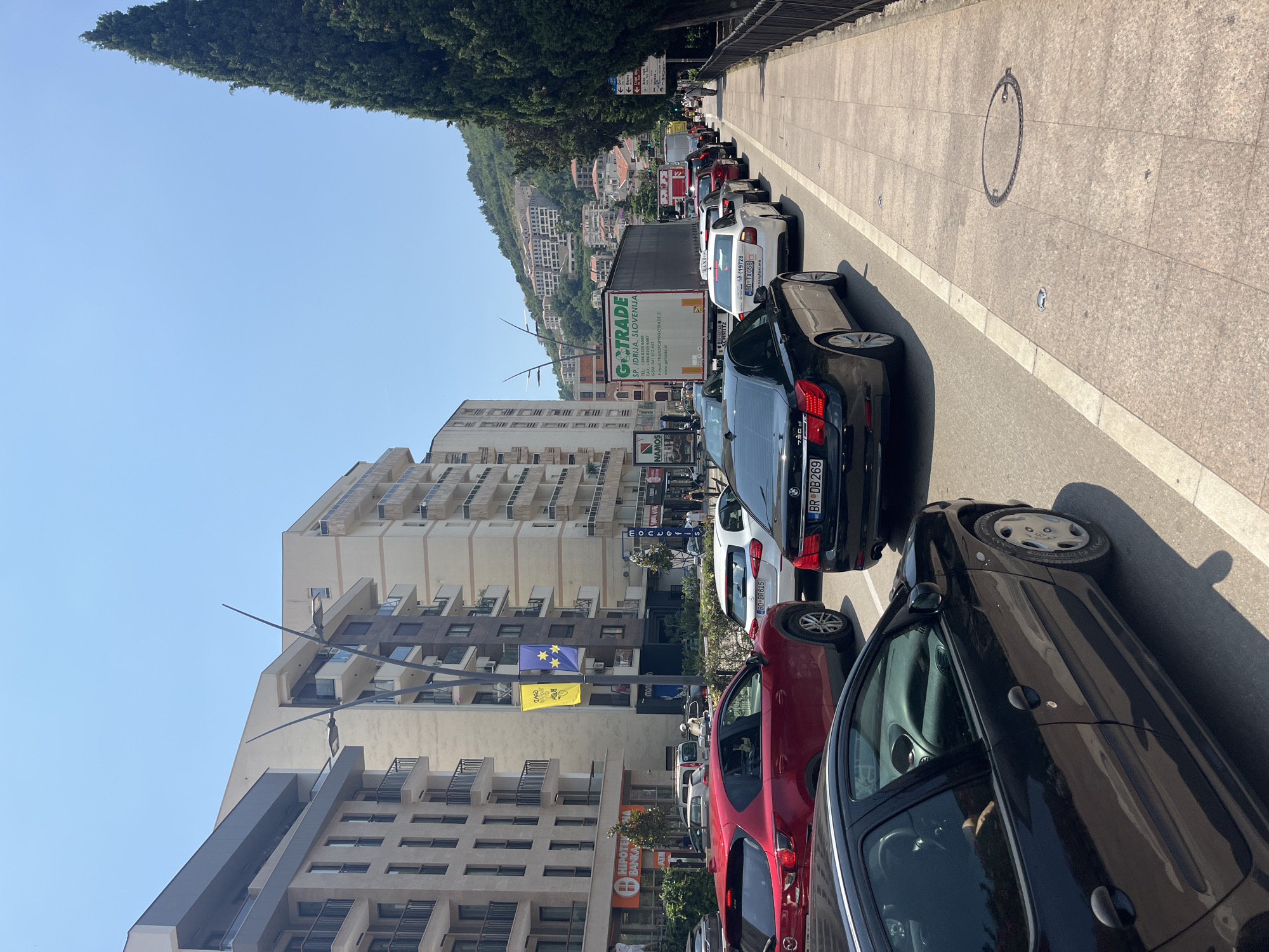 The image captures a street scene in a city, likely during daytime under a clear blue sky. The foreground features a black car on the left, partially obscuring a red car in front of it. To the right, a line of cars is visible, including a black BMW and a white sedan, indicating traffic. Beyond the cars, a sidewalk runs along a row of modern buildings with balconies. A large white truck with "GT-TRADE SP. IDRIJA, SLOVENIJA" text is prominent in the mid-ground, also part of the traffic congestion. A tall evergreen tree stands on the right side of the sidewalk, and further in the background, a hillside is dotted with buildings, suggesting a developed urban area. Pedestrians are sparsely visible on the sidewalk. Two banners, one yellow and one blue with yellow stars, hang vertically from a lamppost in front of one of the buildings. The overall impression is of a busy urban environment with moderate traffic on a sunny day.