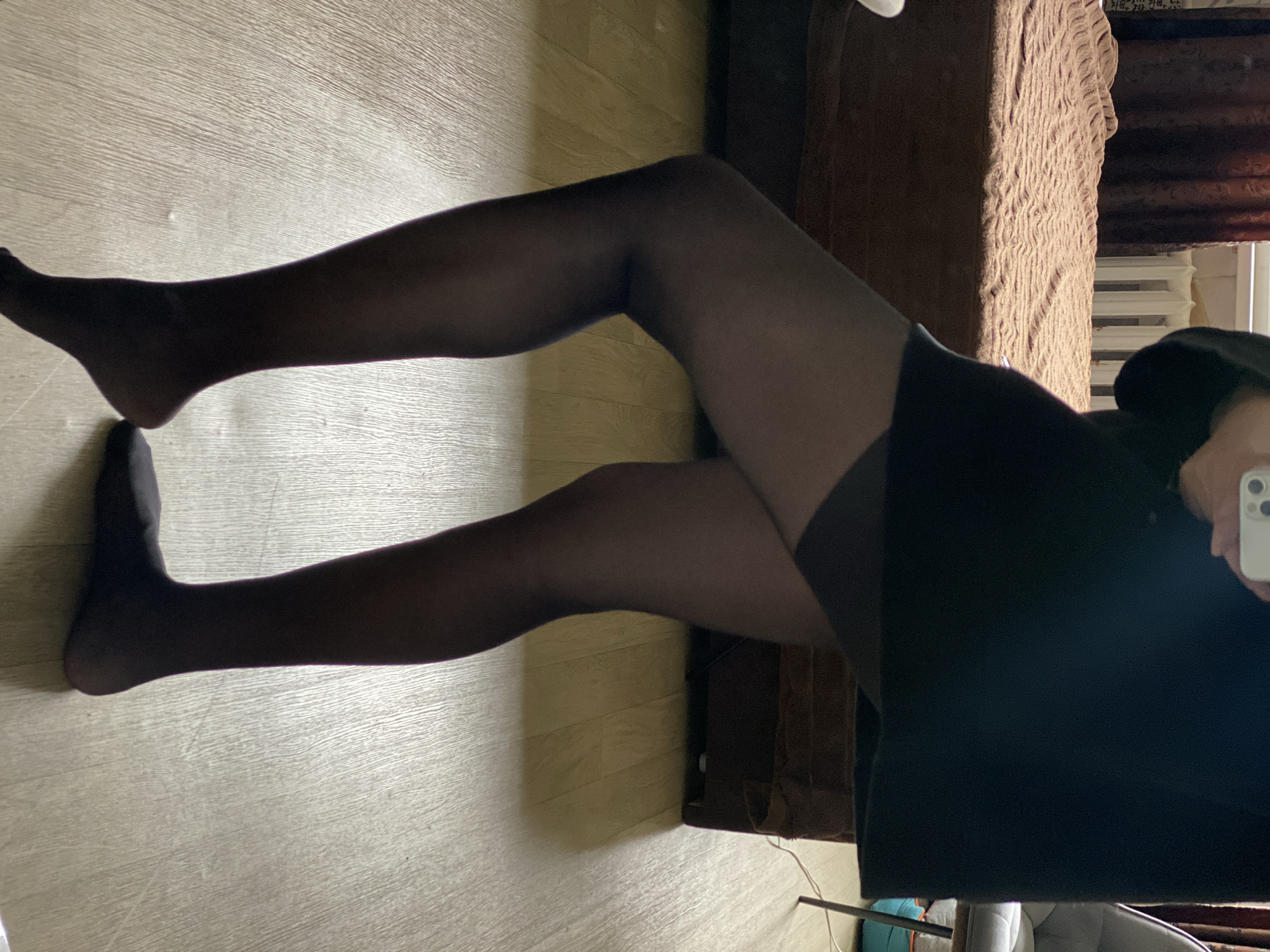 A person wearing black tights and a black dress is lying on a light wood-colored floor. One leg is bent at the knee, the other is straight. The person is holding a smartphone in their right hand, possibly taking a picture. Part of a brown sofa is visible in the background, with a white radiator behind it. The setting appears to be indoors, possibly a living room. There's no evidence suggesting a specific location in Novokuznetsk, Russia.