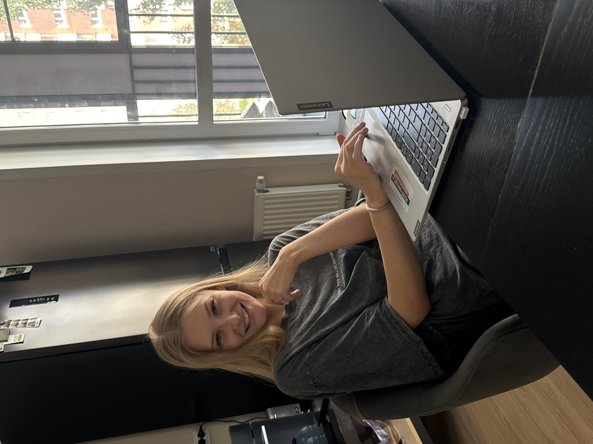A young blonde woman with fair skin and blue eyes is seated at a dark wood table, looking towards the camera and smiling. She is wearing a dark grey t-shirt with faded text on it. Her right hand is resting on the table near her chin, and her left hand is on a silver Lenovo laptop keyboard. The laptop is open and positioned on the table in front of her. To the left of the woman, a tall, black refrigerator is visible, with some papers and magnets attached to its door. Behind her, a window with a white frame looks out onto an urban landscape with a brick building and trees. A white radiator is mounted on the wall beneath the window. The scene appears to be an indoor residential setting, possibly an apartment in the city of Perm, Russia.