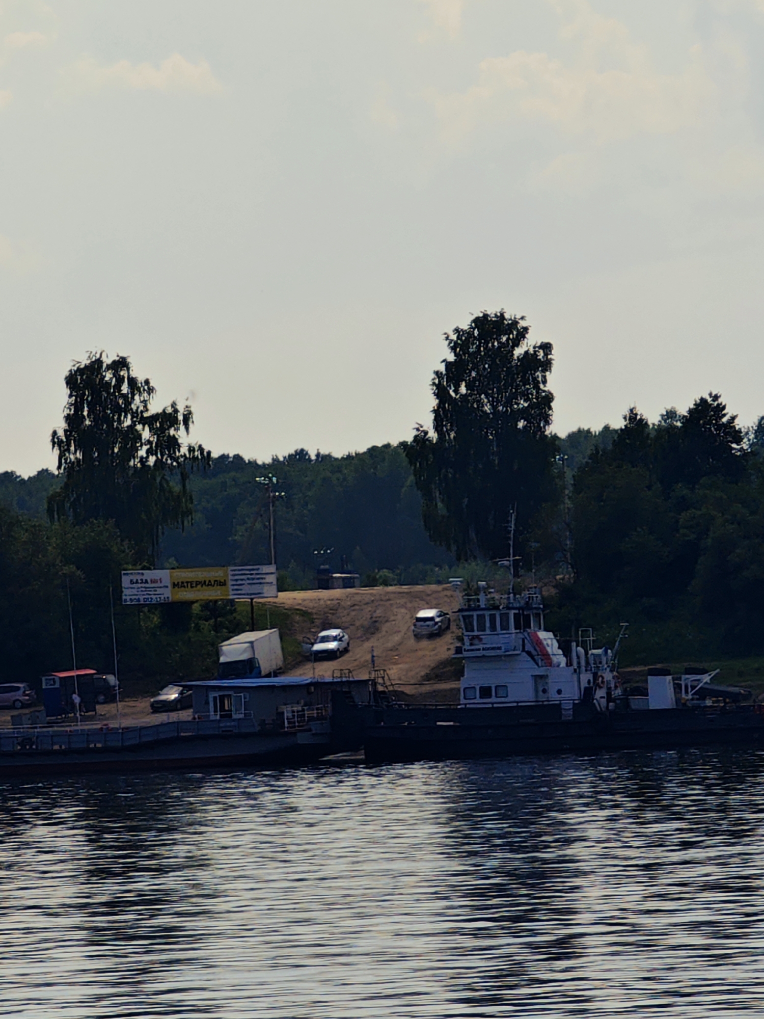 The image depicts a waterfront scene in Erkalovo, Russia. A tugboat is moored to a ferry, which is partially submerged in the water. The ferry is loaded with what appears to be building materials, including a white truck and a blue modular building. In the background, a dirt ramp leads up to a wooded area, and several vehicles are parked near a sign advertising "MATERIALS". The sky is overcast, with scattered clouds. The overall atmosphere is one of industrial activity and transportation.
