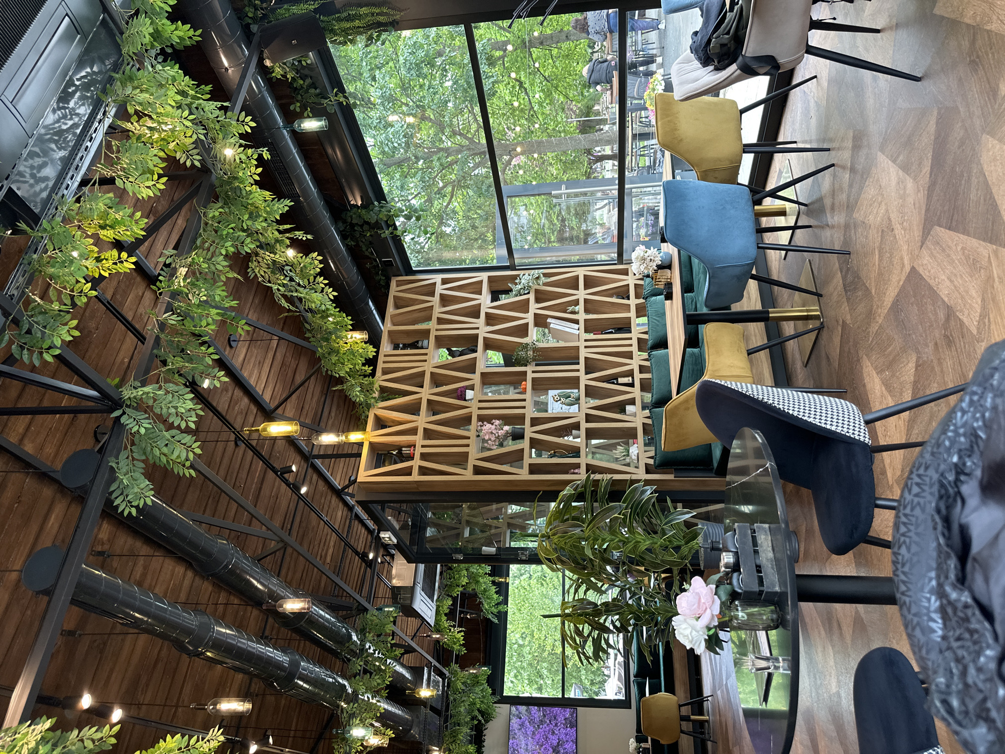 The image shows the interior of a restaurant or cafe. The location is inferred to be Montana, Bulgaria based on prior knowledge. The space has a modern, industrial-chic design with a high ceiling featuring exposed wooden beams, black metal piping, and artificial greenery strung along the pipes. There are pendant lights hanging from the ceiling.
A large window with a view of trees outside is present. A wooden shelving unit with a geometric design is positioned near the window, displaying decorative items such as plants, flowers, and bottles.
The seating area consists of a dark table surrounded by chairs with various upholstery colors (blue, yellow, green, and black/white patterned). Some chairs are unoccupied, while others appear to have jackets or bags on them, suggesting patrons may be present. There are fresh flowers in a vase on the table.
The floor has a geometric wood pattern. Overall, the atmosphere is bright and airy due to the natural light from the large window and the abundant greenery.