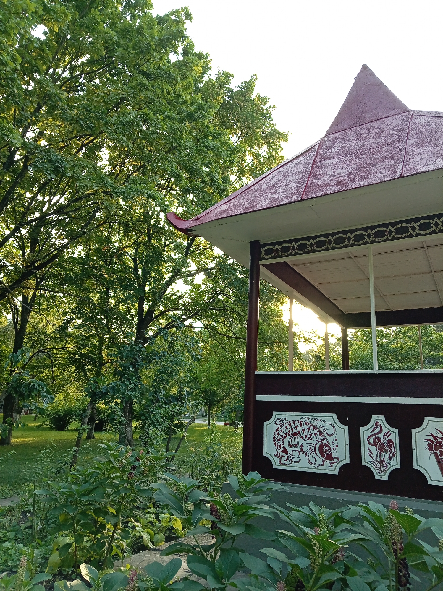 The image shows a partially visible, ornate gazebo in Yessentuki, Russia. The gazebo has a red, multi-peaked roof and dark red siding adorned with white and red decorative panels. One panel clearly depicts a stylized red fish on a white background, while another shows a red crane. The gazebo is situated in a lush, green park setting with tall trees and various plants in the foreground. Sunlight filters through the trees and the open structure of the gazebo, creating a warm glow. The scene is captured from a low angle, emphasizing the foliage and the detailed craftsmanship of the gazebo.