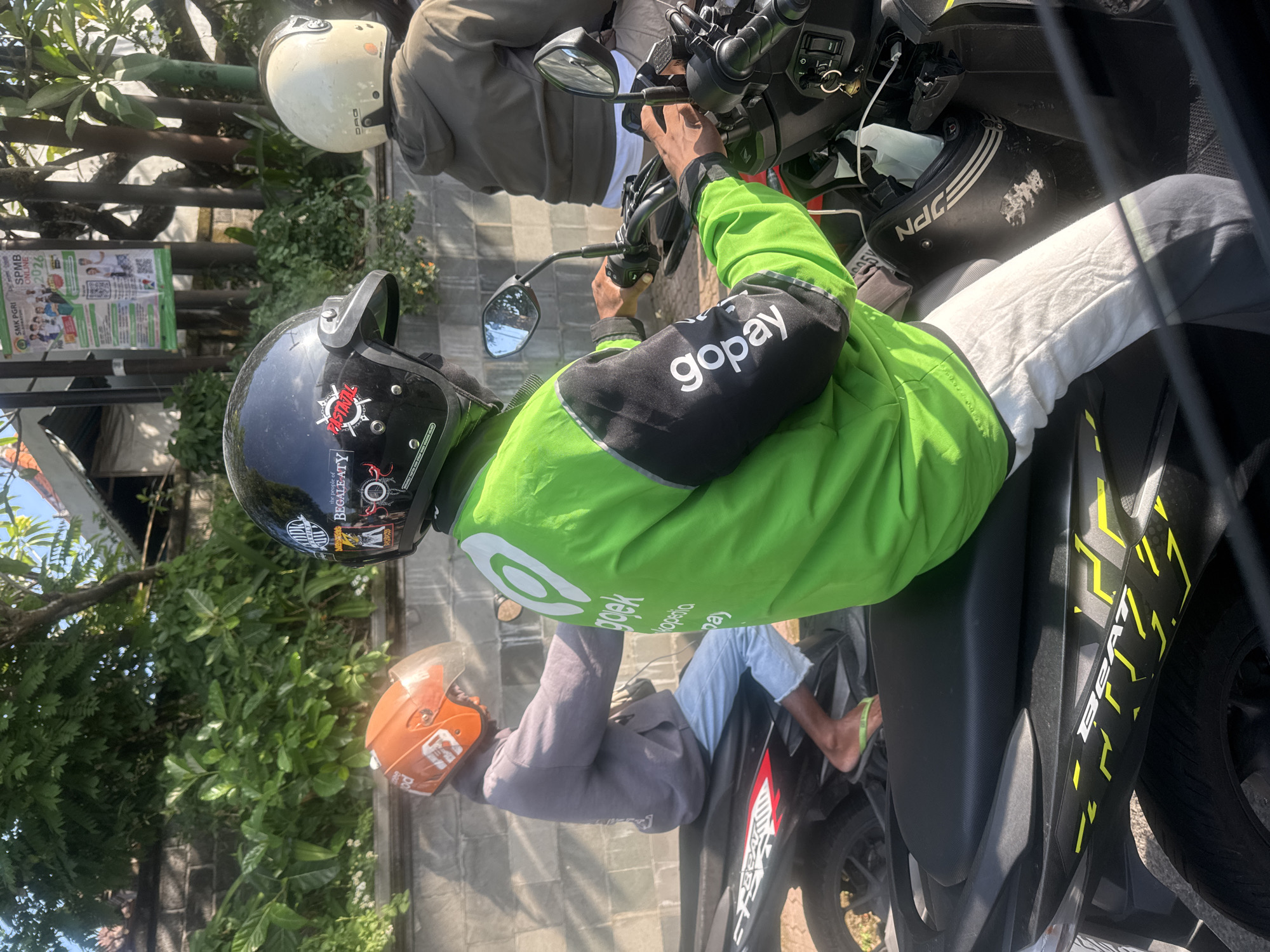 The image shows several people on motorcycles in Sukawati, Indonesia. One person is wearing a bright green jacket with "gopay" printed on the sleeve, indicating they may be a driver for a ride-hailing service. Another person has an orange helmet, and another a white helmet. There is a parked motorcycle with "BEAT" written on its side, and the parked motorbikes have riders onboard. There is a sign for SMK PGRI showing 2026. The scene appears to be a busy street, with trees and foliage in the background.