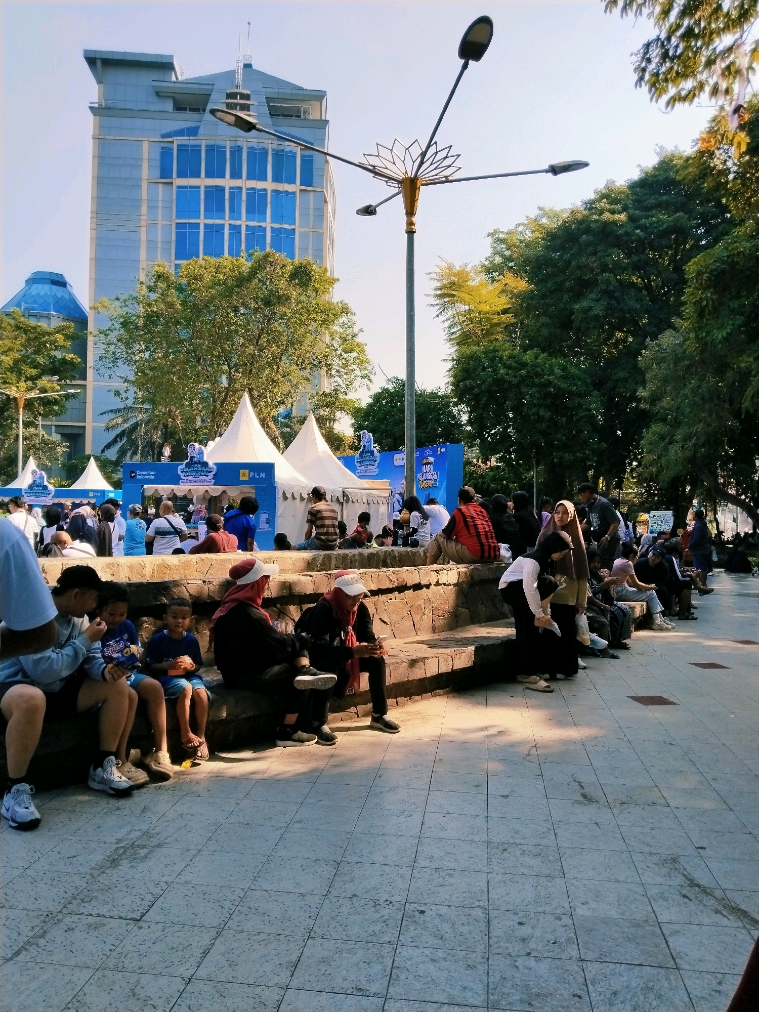 The image depicts an outdoor event or gathering taking place in front of a modern glass-fronted building, likely in an urban park setting. The building in the background features a distinctive blue hue and appears to be a corporate office or administrative center. In the middle ground, several white tents with pointed roofs are set up, suggesting a fair, festival, or exhibition. Banners with Indonesian text and logos, including "PLN," are visible on and around the tents, indicating a possible connection to the state electricity company.
A diverse group of people are present. In the foreground, several individuals, including children and adults, are seated on tiered stone steps. Some are casually dressed, while others wear headscarves. They appear to be watching or participating in the event. Further back, a larger crowd is gathered around the tents, suggesting active engagement with the activities or vendors. The environment is sunny with clear skies, and mature trees are present, providing shade and a natural backdrop to the scene. The ground is paved with patterned tiles. The overall impression is a lively community event in an urban park, possibly during a daytime celebration or promotional activity. The presence of the PLN branding and the urban park setting strongly suggests this location is the Taman Surya Boulevard, a public space often used for events in Surabaya, Indonesia, adjacent to the PLN building.