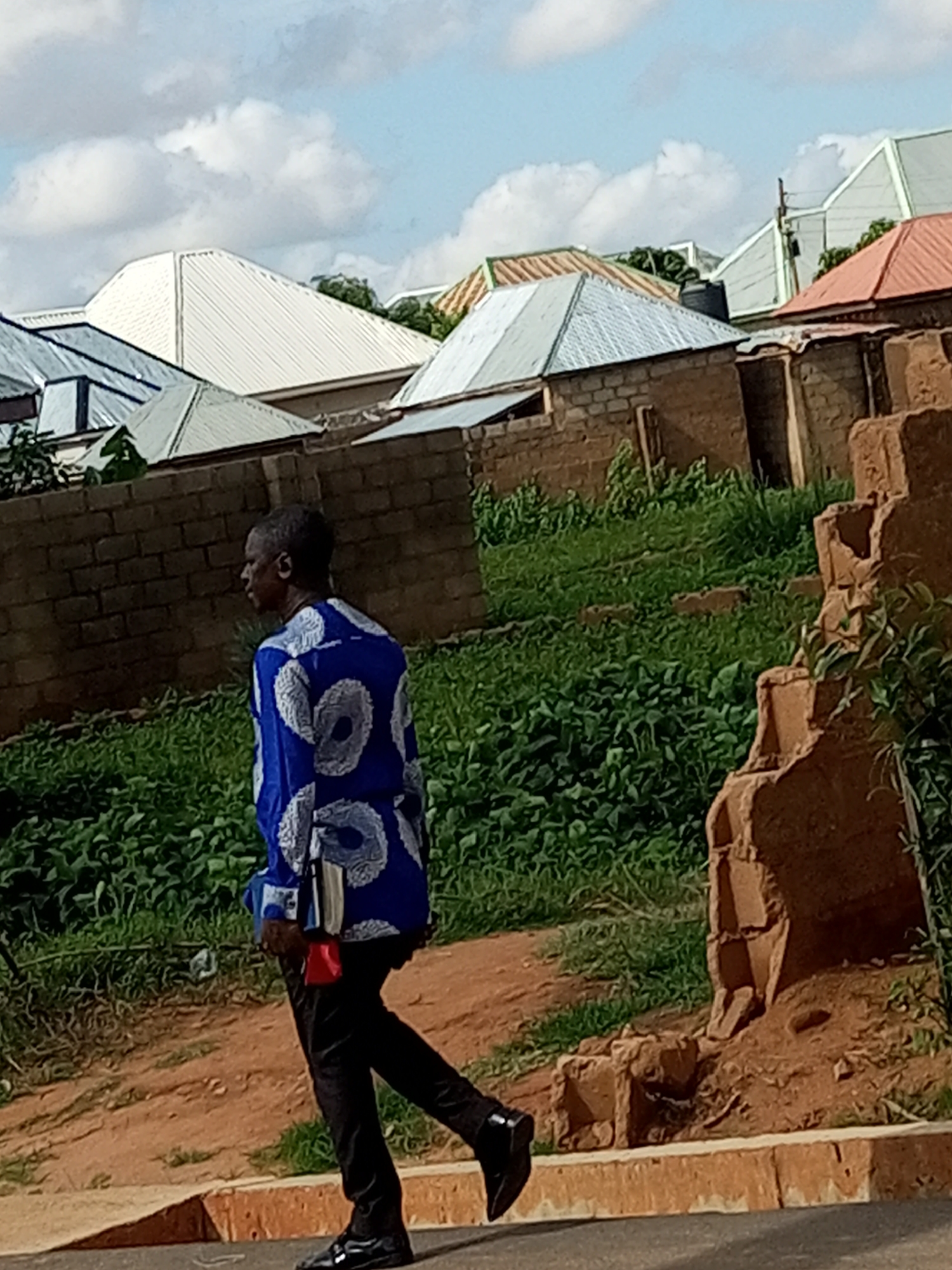 The image depicts a man walking on a paved surface in Gombe, Nigeria. He is wearing a blue shirt with a white circular pattern and black pants. He appears to be carrying a red and white object attached to his side. The background shows a brick wall, lush green vegetation, and several buildings with corrugated metal roofs of various colors, suggesting a residential or urban area. The sky is visible with scattered white clouds. The scene is captured outdoors during daylight.