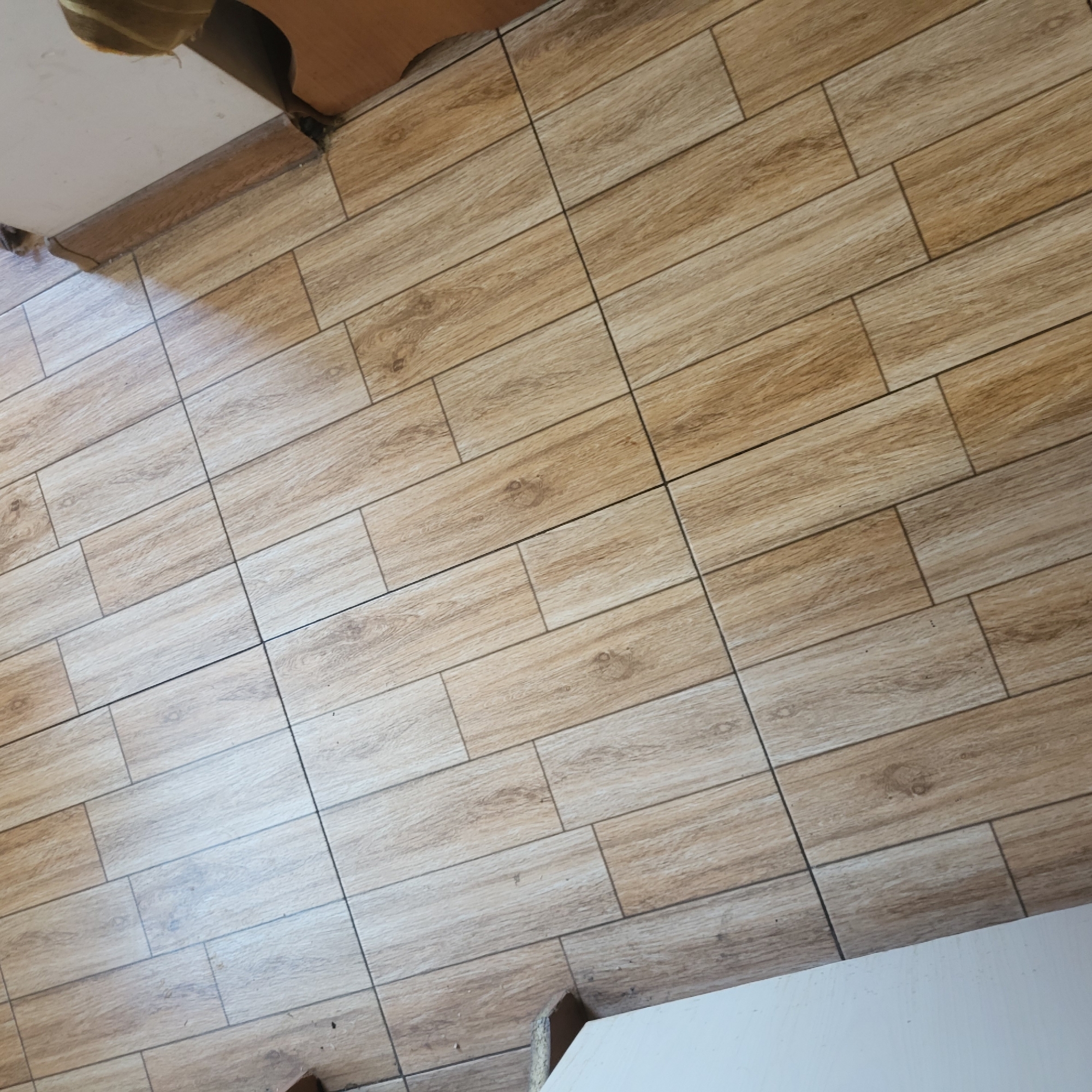 The image presents an indoor floor covered with rectangular ceramic or porcelain tiles designed to resemble light brown wood planks. The tiles exhibit varied grain patterns and shades of beige and light brown, with some featuring darker, knot-like details, mimicking the texture of natural timber. They are laid in a staggered, offset pattern, separated by thin, dark gray grout lines.
Natural light illuminates the floor, casting a noticeable shadow across the upper left section of the frame, indicating a daytime setting. In the top left corner, a white ceiling or wall meets a wooden trim, with some visible imperfections or debris. The bottom right corner reveals a white surface, possibly a wall or the base of furniture, extending into the frame. No people or specific activities are depicted. The scene is a static view of the flooring within a room in Yerevan, Armenia. No visible text is present.