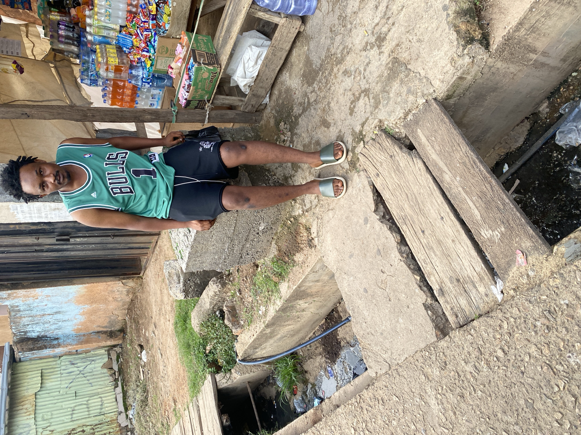 In Jos, Nigeria, a young man stands beside a roadside stall. He wears a green Chicago Bulls basketball jersey and black athletic shorts, with green slip-on sandals on his feet. His dreadlocks are visible. The stall is stocked with an assortment of bottled beverages and packaged snacks. Behind the man, a corrugated metal fence marks the boundary of a property. In the foreground, a concrete channel, likely a drainage system, runs alongside a wooden walkway. The channel contains murky water and debris, indicating a potentially unhygienic environment. The overall scene depicts a common urban setting in Nigeria, characterized by modest infrastructure and commercial activity.