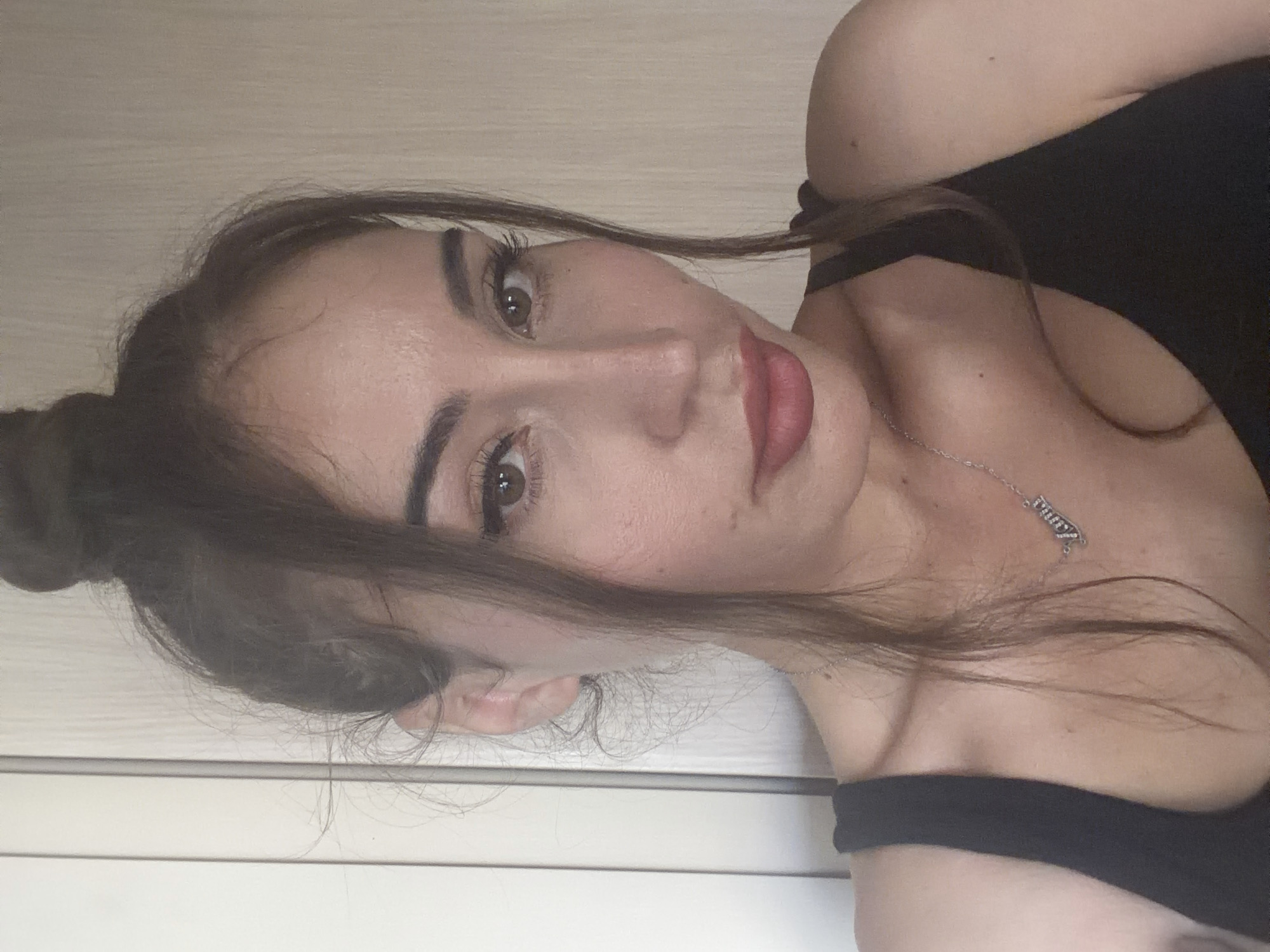 A medium shot, chest-up, indoor portrait of a young adult female with her hair pulled up in a bun. She is wearing a black tank top and a silver necklace with a nameplate. The background consists of a beige wall on the right and a light-colored door on the left. The image appears to have been taken in Tersefanou, Cyprus.