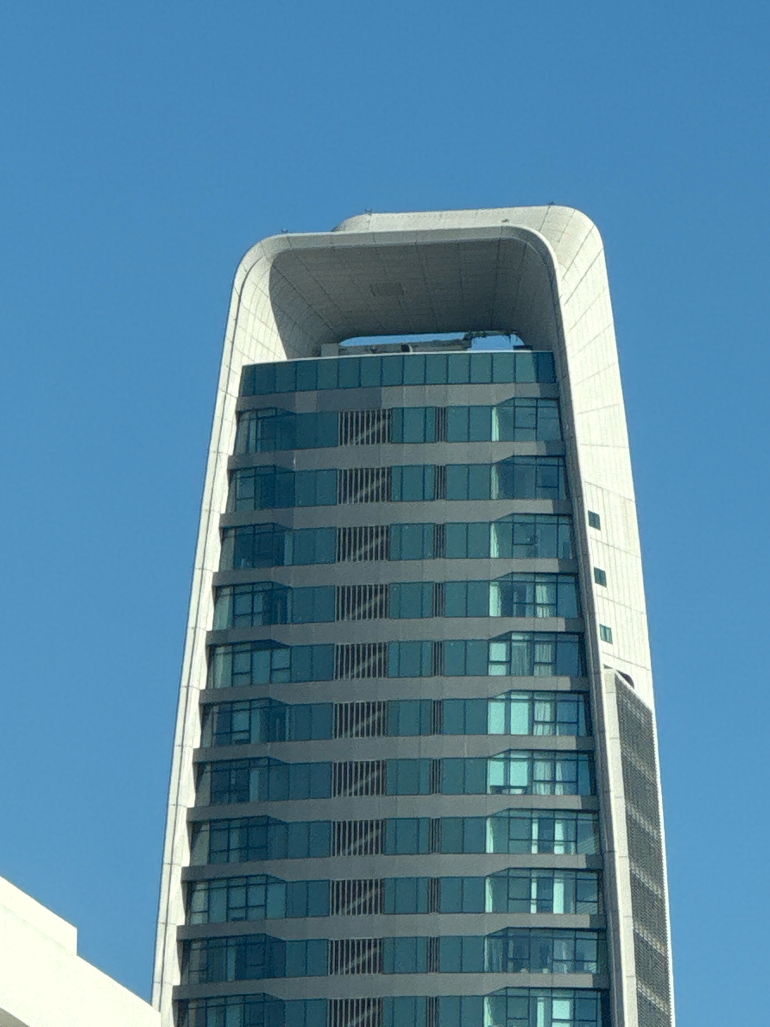 A modern, glass-paneled skyscraper with a distinctive U-shaped opening at its crown stands against a clear blue sky. The building's facade is composed of horizontal bands of blue-tinted windows interspersed with gray structural elements. The skyscraper's base is not visible in this close-up view, but a white, angular structure is partially visible in the lower left corner, suggesting it is part of a larger complex. The architecture implies a contemporary urban setting, possibly in Limassol, Cyprus, known for its modern developments. No people or discernible actions are visible.