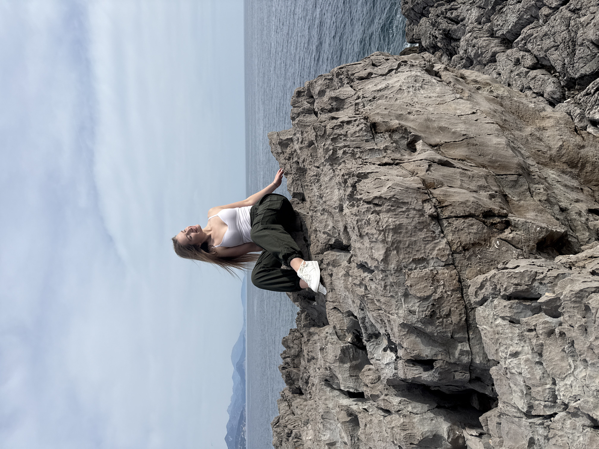 A woman is posing on a rocky cliff in Boreti, Montenegro. She is wearing a white tank top, dark green pants, and white sneakers. The cliff is gray and textured, with the Adriatic Sea and coastline visible in the background. The sky is partly cloudy. The woman is positioned on the cliff's edge, with one hand touching the rock face. She is looking upward and smiling.