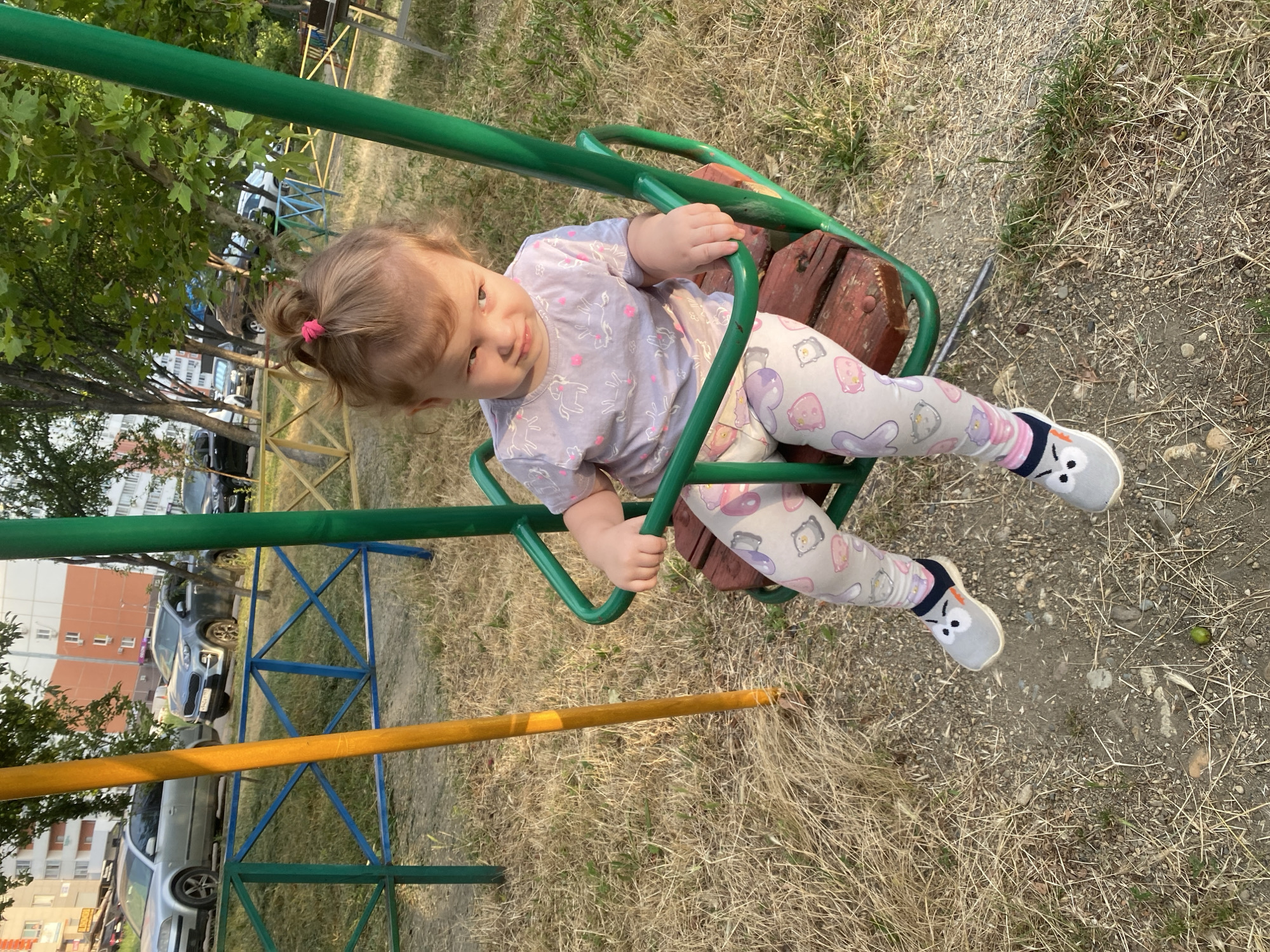 A young child, likely a toddler, is seated on a wooden swing. The child has light brown hair pulled into a small pink ponytail and is wearing a light purple t-shirt with a unicorn pattern and light-colored leggings adorned with various cartoon characters, including what appear to be animals. The child is also wearing light gray slip-on shoes with cartoon faces on them and striped socks. The child is holding onto the green metal frame of the swing. The swing set appears to be made of green metal and has a wooden seat. The scene is set outdoors, with dry grass and dirt covering the ground. In the background, there are trees, a metal fence, and buildings, including a multi-story apartment building. Cars are visible parked in front of the buildings. The lighting suggests it is daytime, likely late afternoon due to the long shadows. The child's expression is one of gentle curiosity or amusement.