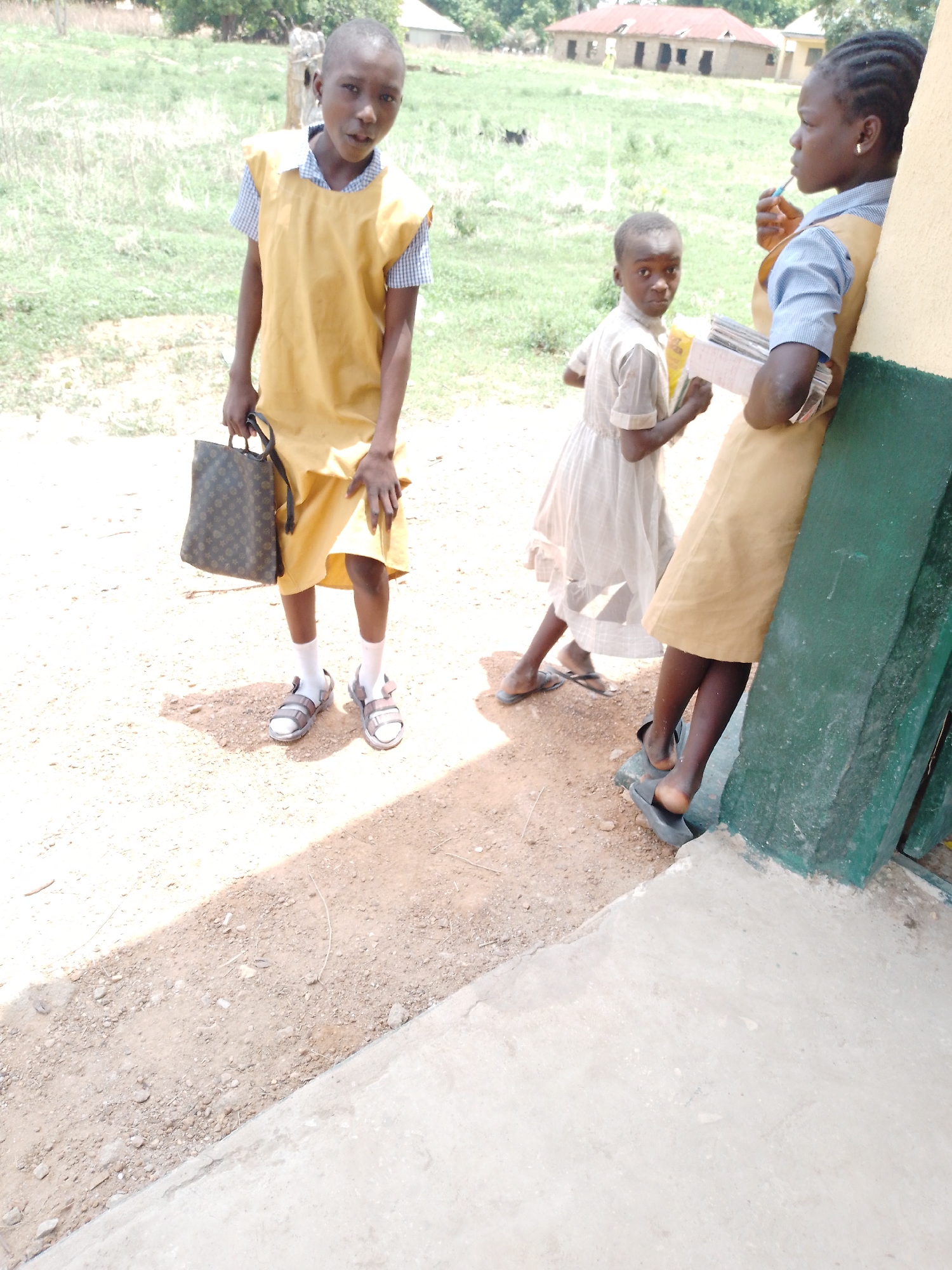Three young girls stand in an outdoor setting, their clothing suggesting they are students in a school. The foreground consists of a dirt and gravel ground with a portion of a concrete structure on the lower right corner. A girl on the left stands out wearing a yellow dress over a blue and white checkered shirt. She carries a handbag. Her legs are visible, clad in white socks and sandals. To the right and in the background stands a girl in a light-colored dress. Further to the right, and slightly behind the girl in the light dress, another young girl stands leaning against a wall. She is wearing a yellow dress over a blue shirt, and has a toothbrush in her mouth. She is also holding several books. In the background are buildings with red roofs and some sparse vegetation. The location of the city is unknown.