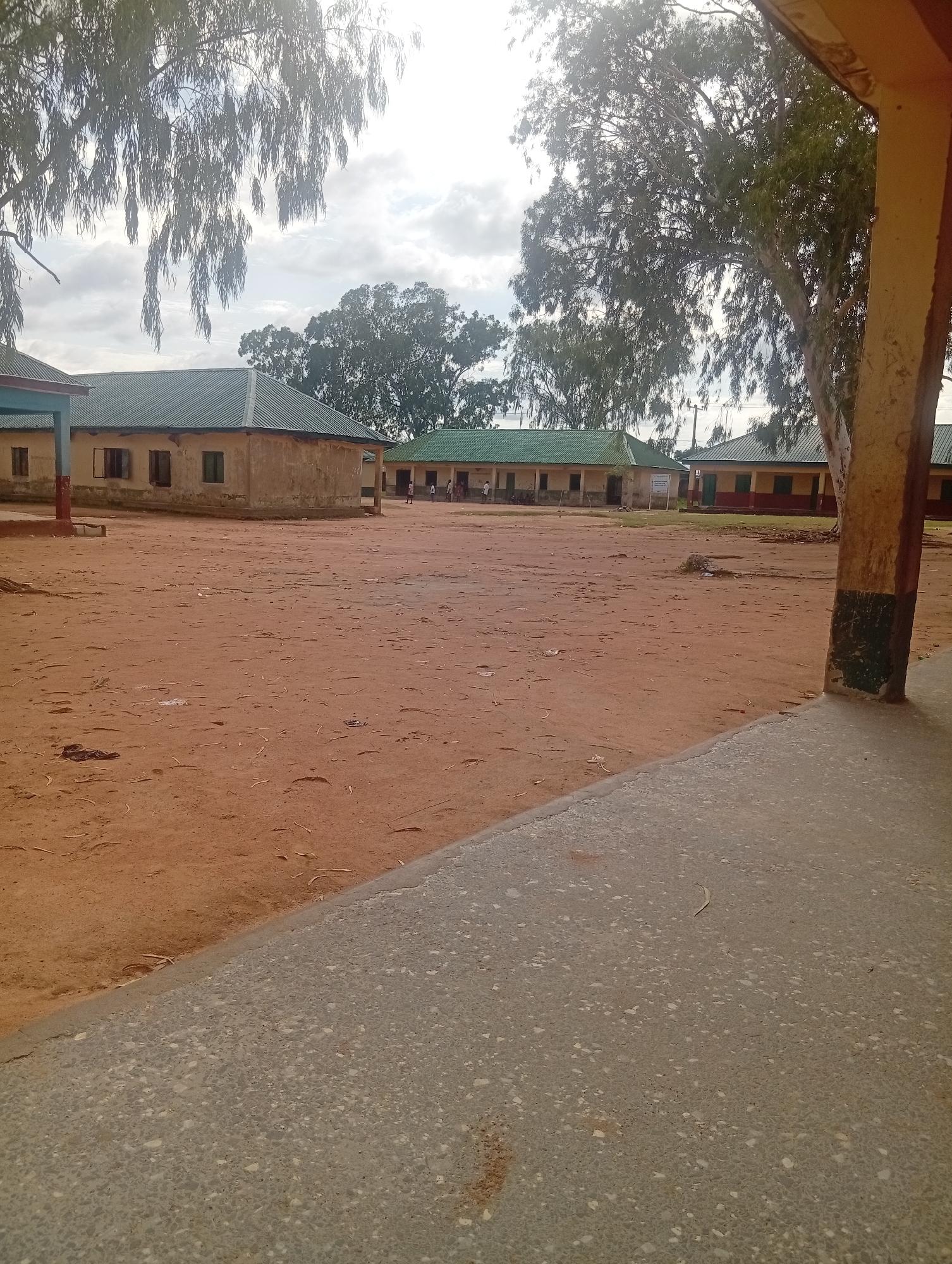 The image depicts an outdoor compound, likely a school, featuring several single-story buildings under an overcast sky. In the foreground, a speckled grey concrete surface occupies the lower-left, diagonally transitioning into an expansive area of reddish-brown dirt. Small debris, including dry leaves and twigs, are scattered across the dirt, with faint footprints discernible.
Several single-story structures with green corrugated metal roofs are visible in the midground. The closest building on the left features light-colored walls, some sections painted pale blue, and multiple rectangular window openings, some appearing shuttered. Further back, another similar building has light-colored walls and a legible, white rectangular sign positioned in front of it. Towards the right, partially obscured by a dark-stained concrete column of the structure from which the image was taken, is a third building with a green roof and walls painted with a darker band (possibly red or maroon) below a lighter section.
In the background, numerous trees with dense green foliage are scattered, their leaves partially obscuring the sky, which shows a bright, diffused light suggesting an overcast or hazy sunny day. Several small, indistinct figures, consistent with people (likely children or adults), are visible near the buildings in the far right background, appearing to be standing or moving around. The overall scene suggests a dry, dusty environment.